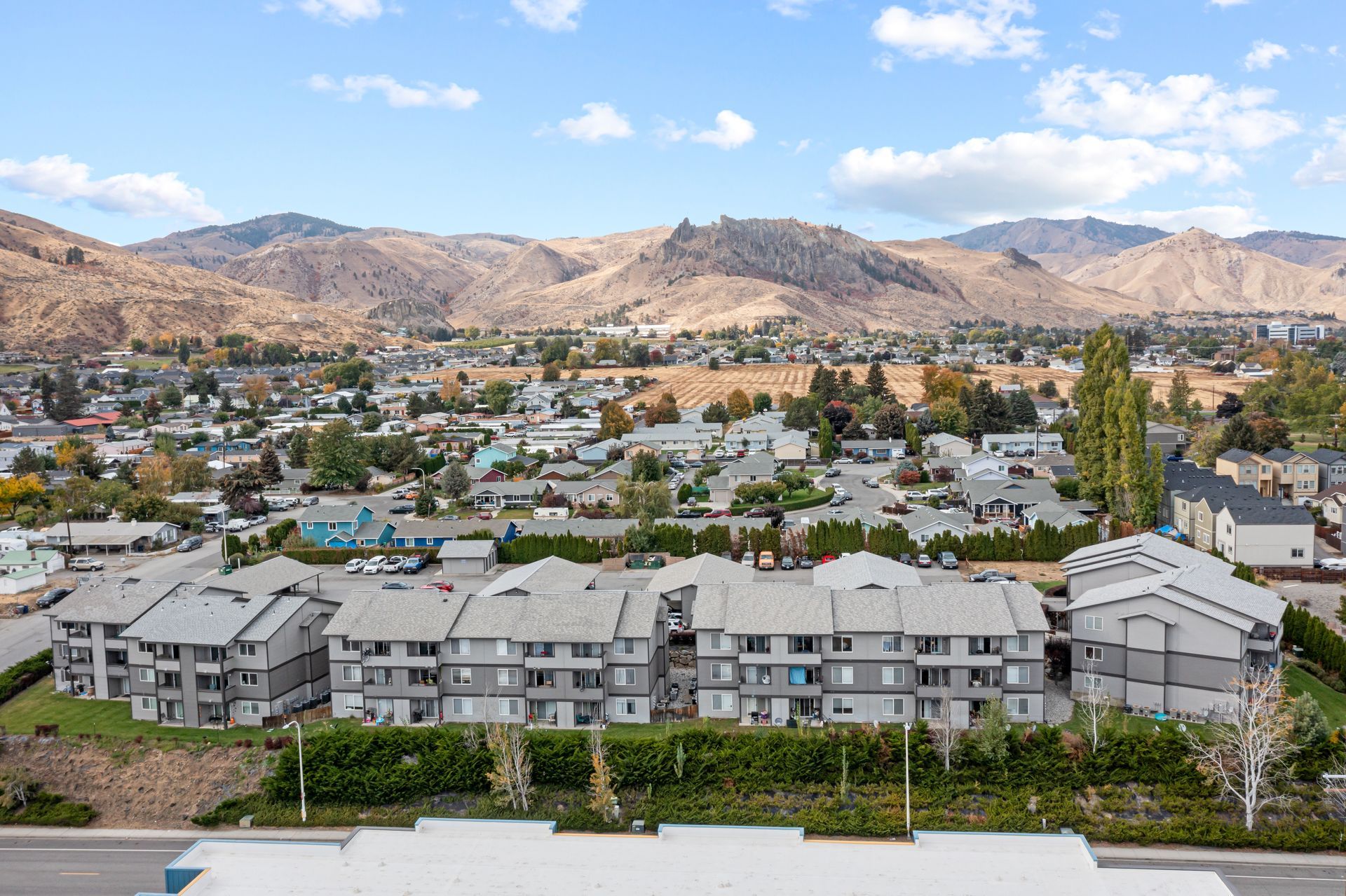Apartment buildings with gray roofs, set against a town and mountain backdrop under a blue sky.