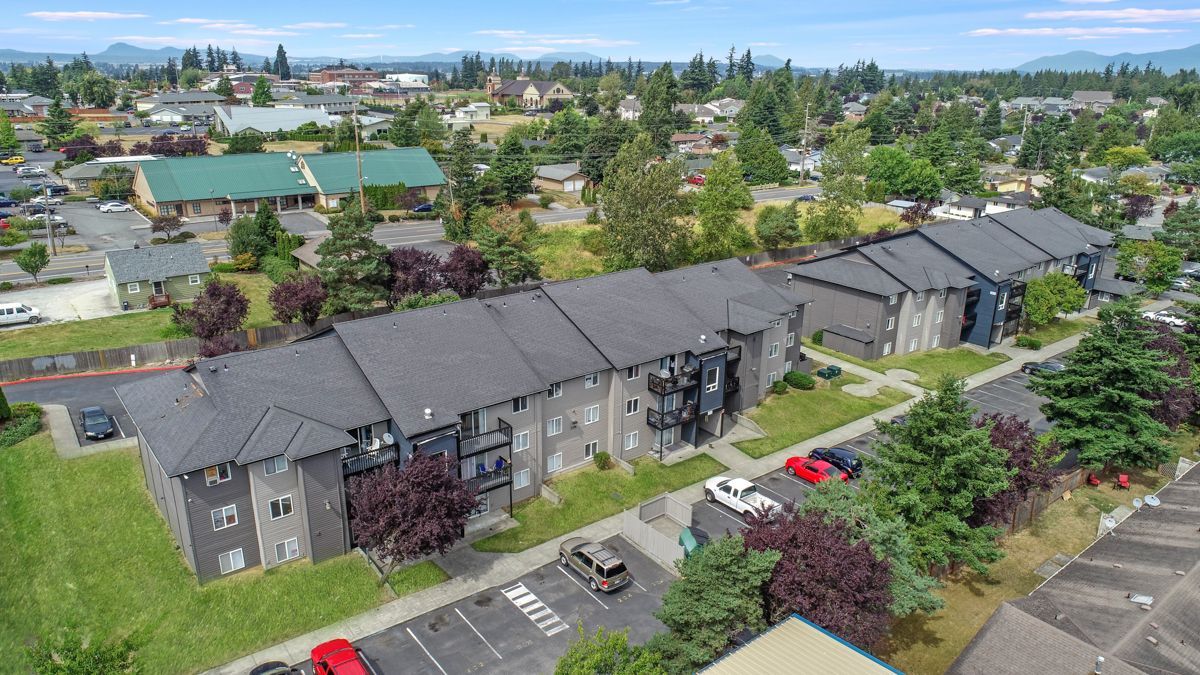 Aerial view of apartment buildings with dark roofs, surrounded by trees and a parking lot.