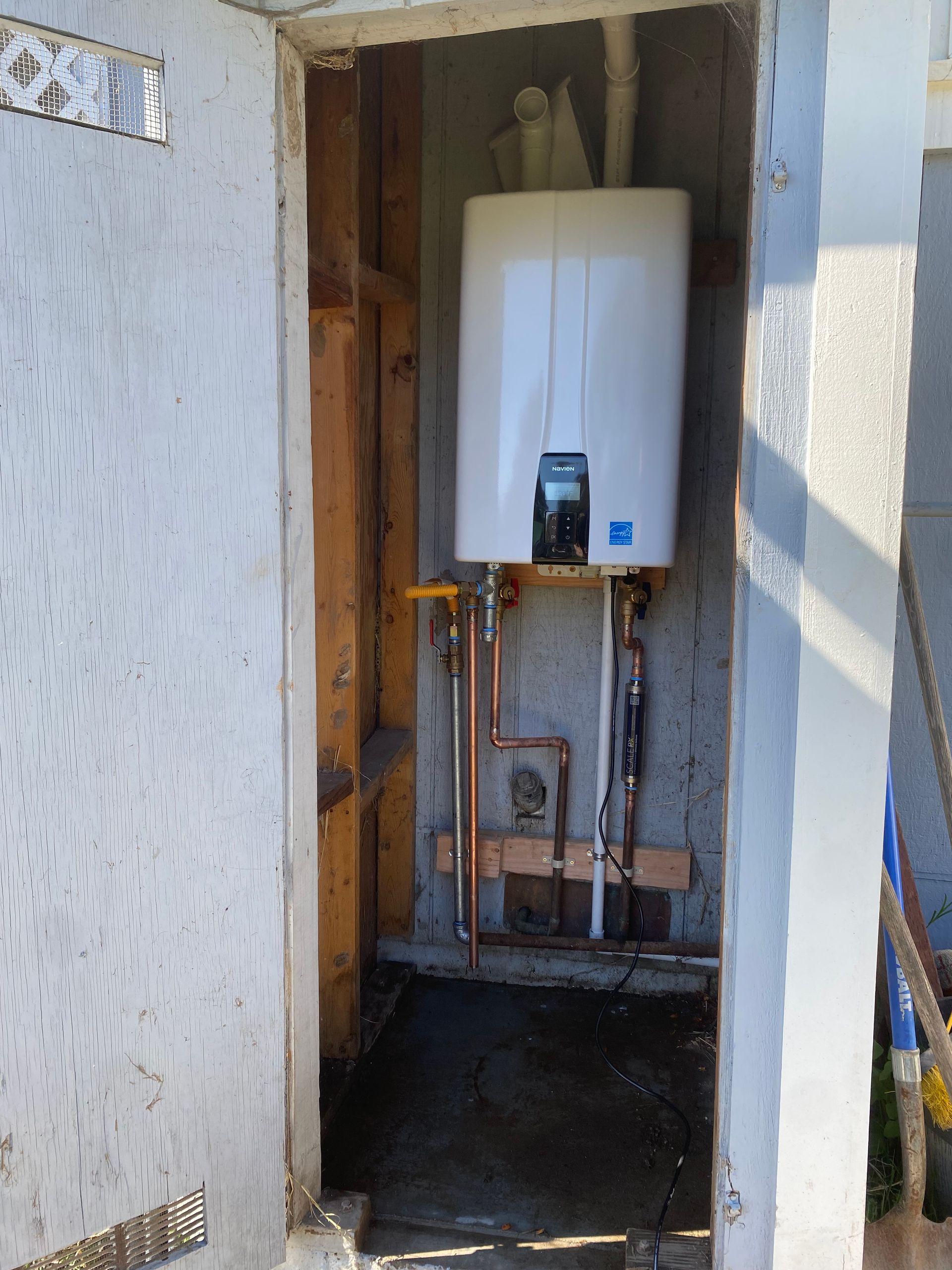 Wall-mounted white boiler in a narrow unfinished utility closet with exposed pipes and wiring.