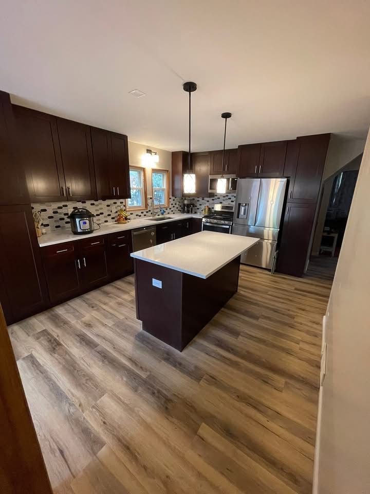 Modern kitchen with dark wood cabinets, island, stainless appliances, and wood-look flooring.