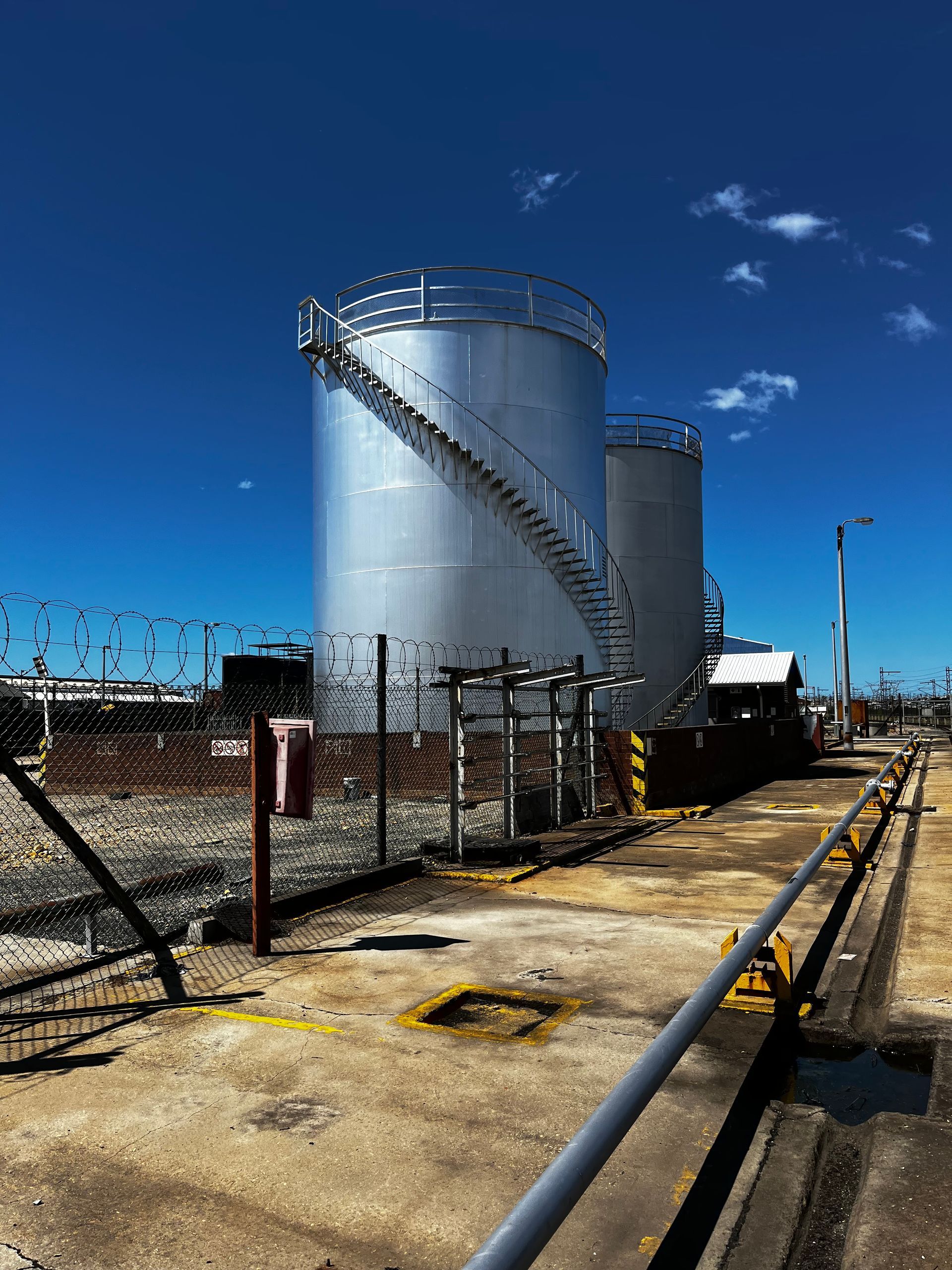 Two silver industrial storage tanks with staircases, behind a chain-link fence, under a blue sky.