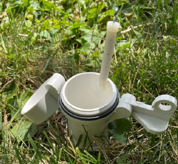 White plastic pool skimmer with a lid, lever, and internal tube, resting in grass.