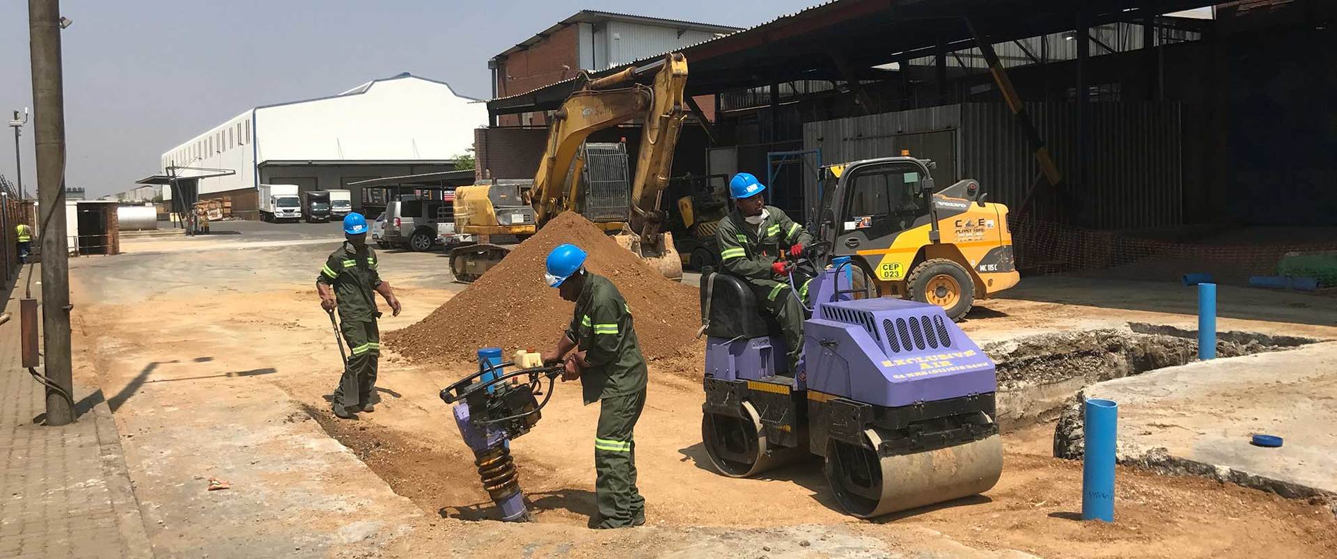 Construction workers operate machinery on a road, including a roller, with a large pile of dirt and a warehouse in the background.