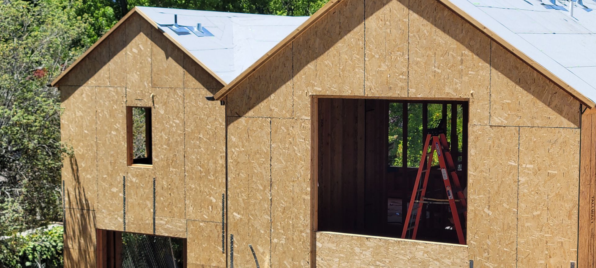 A house under construction, with OSB sheathing and a ladder visible inside a window opening.