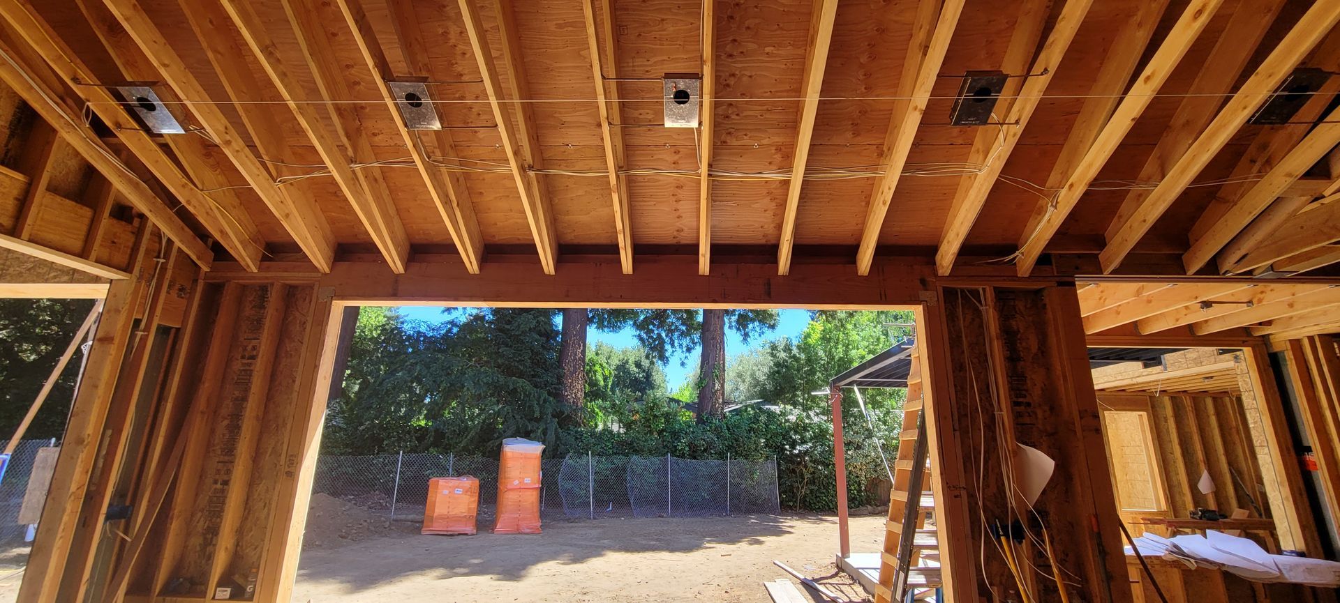Wooden framework of a building's interior, looking out to greenery and sunlight, under construction.