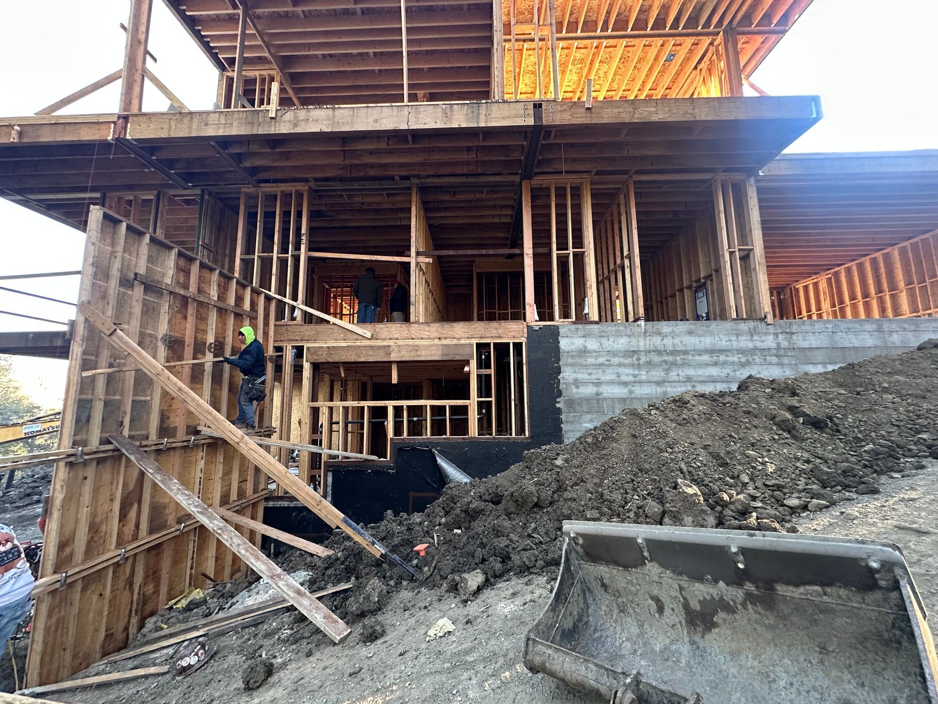 Construction site with wooden framing, excavator, and workers; hillside setting.