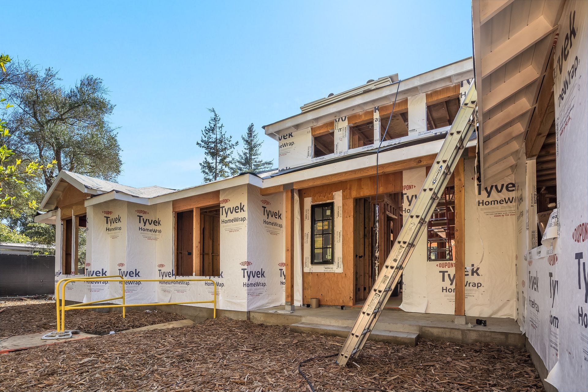 House under construction with Tyvek wrap, wood framing, and a ladder leaning against the facade.