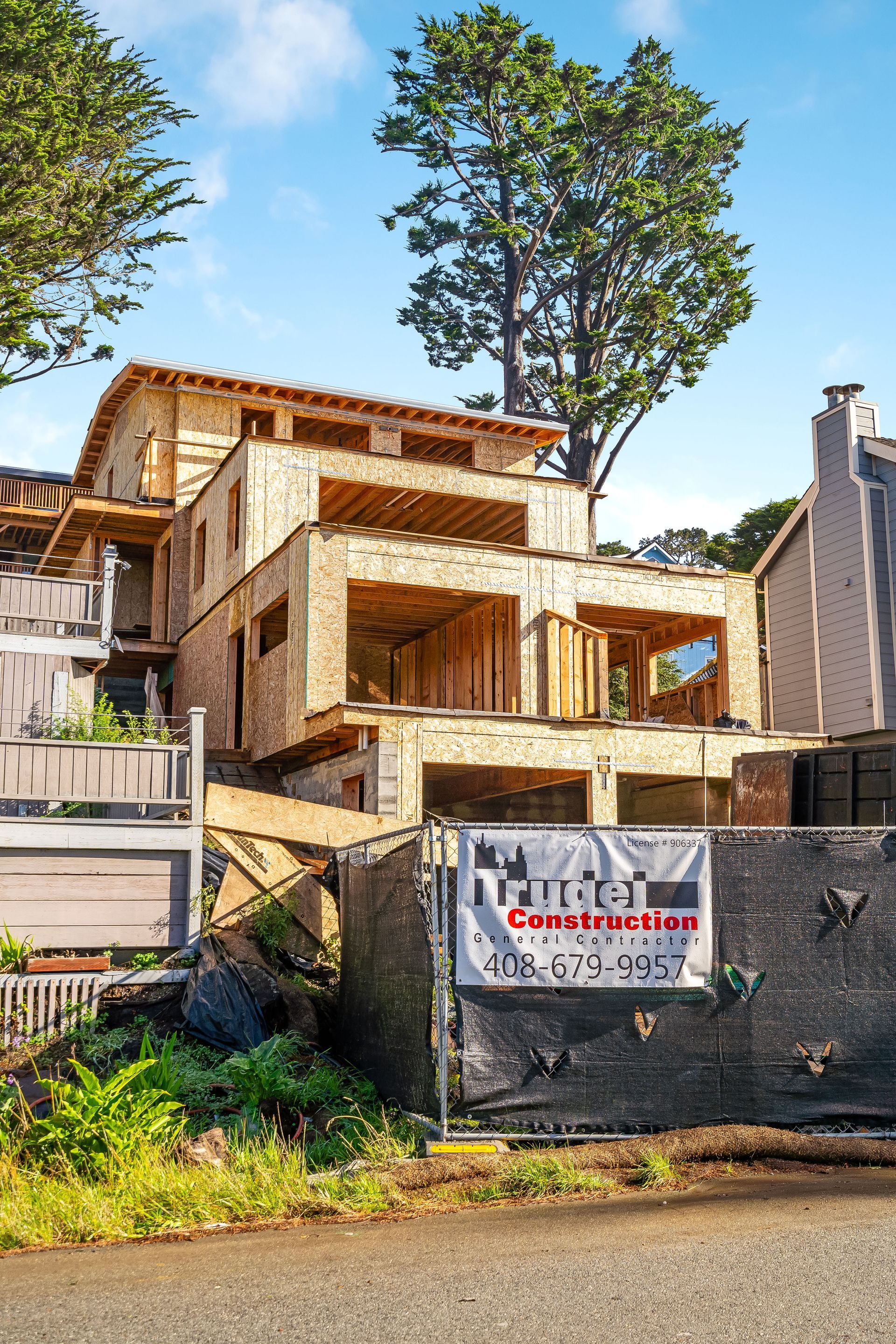 Building under construction, wood frame structure, surrounded by a black fence and greenery.