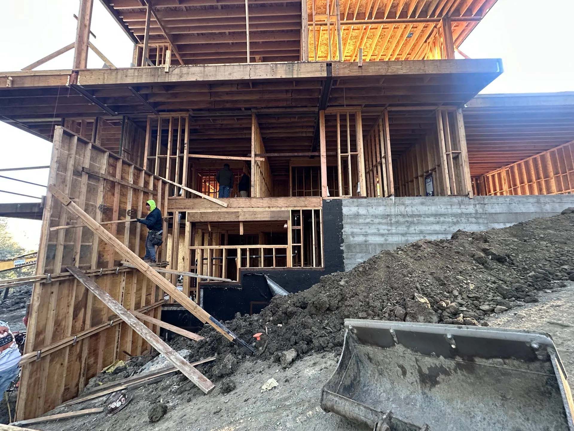 Wooden frame of a house under construction on a concrete foundation, lumber scattered on the ground.