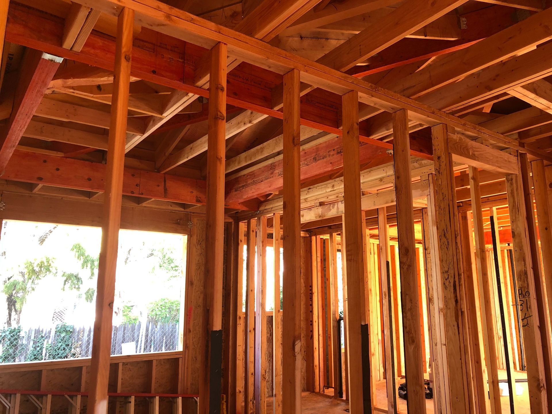 Interior view of a building under construction, showing wooden studs, rafters, and a window opening to the outside.