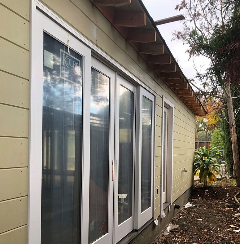 Exterior of a building with white-framed glass doors and siding. A tree is to the right.