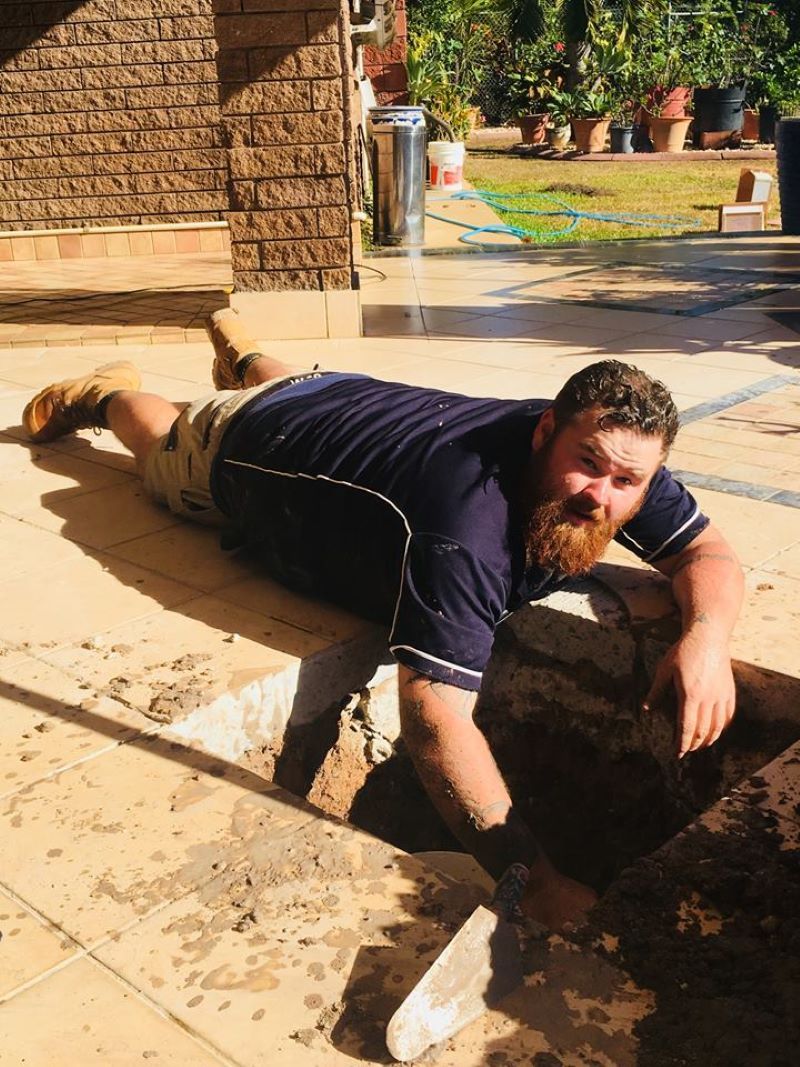 A Man With a Beard is Laying on the Ground Digging a Hole With a Shovel — Absolute Plumbing Australia in Howard Spring, NT
