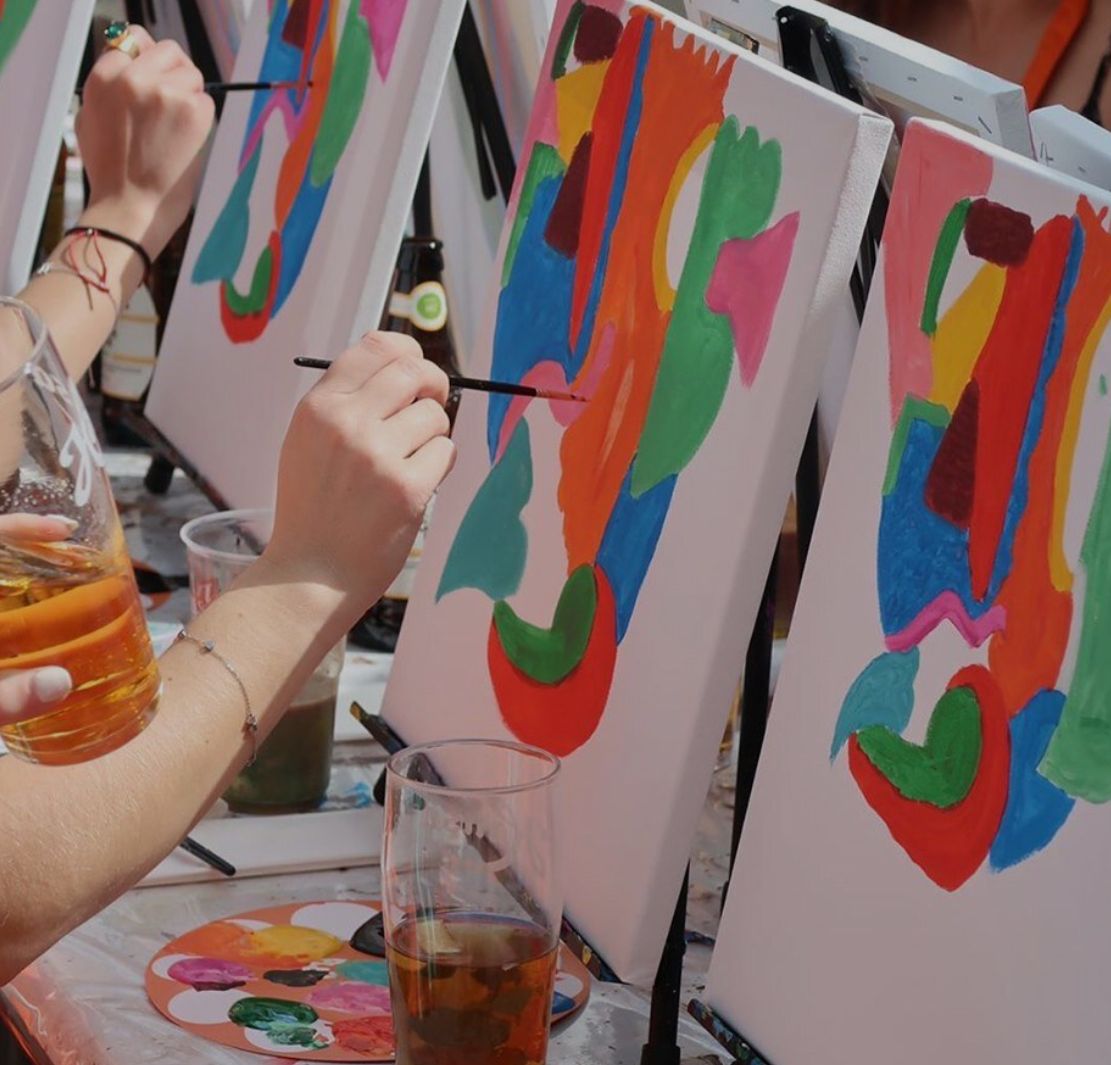 People painting with colorful abstract designs on easels, drinks present.