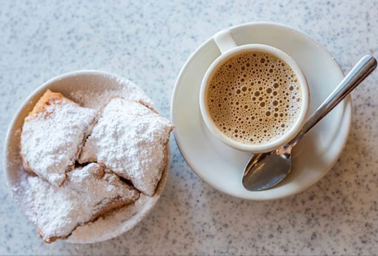 Beignets dusted with powdered sugar and coffee in a white cup on a saucer, with a spoon.