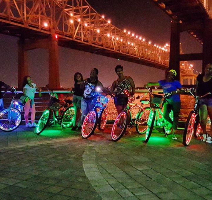 Group of people with lit-up bicycles pose at night under a bridge with city lights.