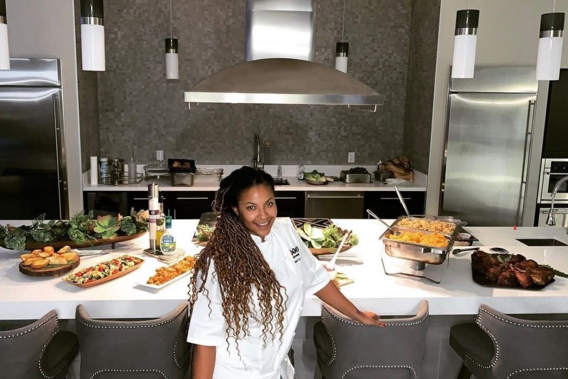 Chef standing in a modern kitchen with food displayed on the counter; smiling.
