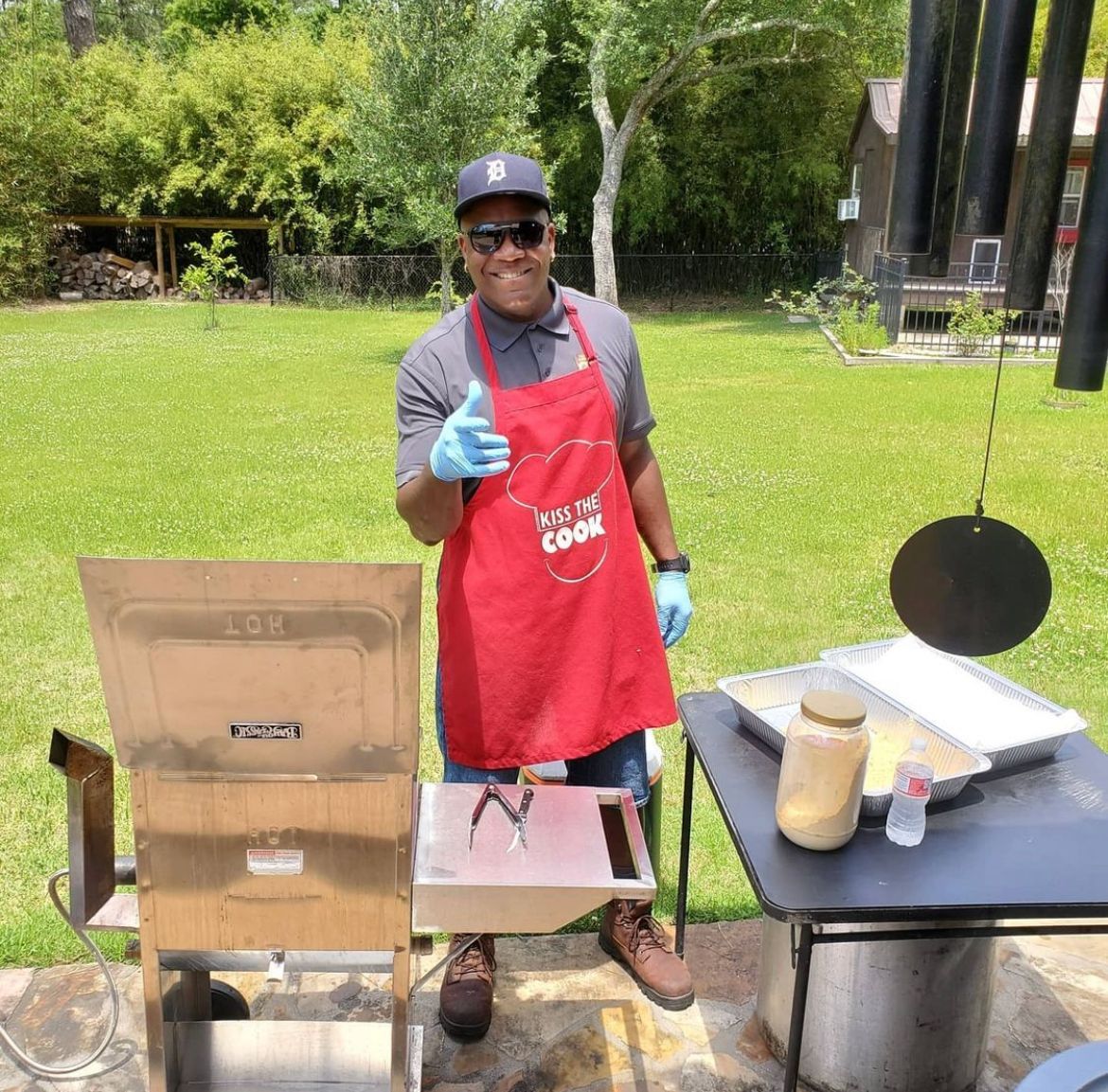 Man grilling outdoors, wearing apron, giving thumbs up. Sunny day, green grass, and cooking equipment are visible.