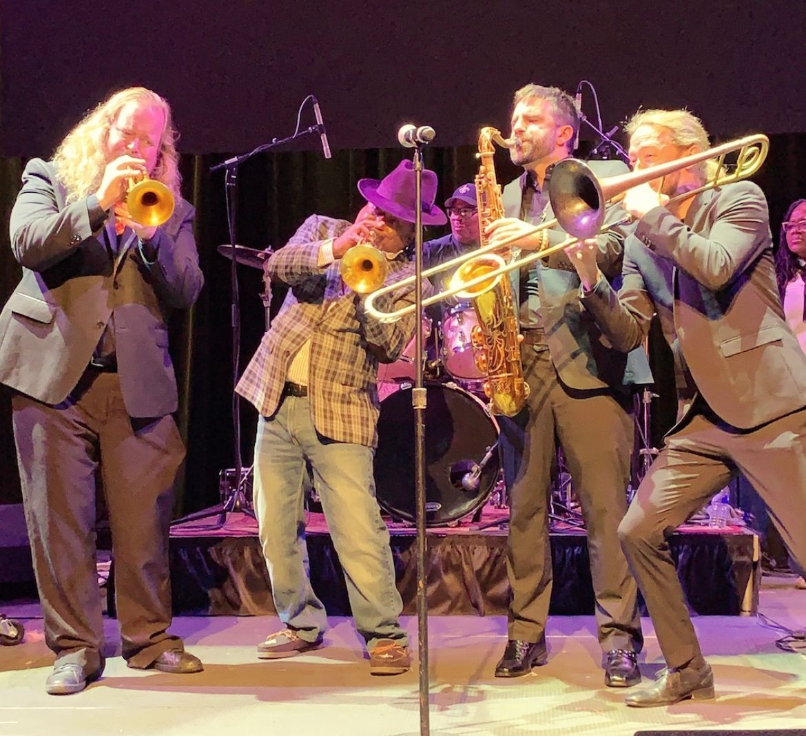 Band on stage: trumpet, trombone, sax, and trumpet players performing. Stage lights illuminate.