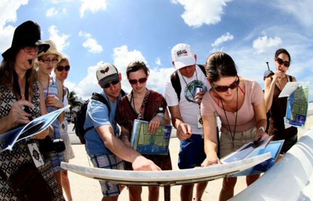 A group of tourists consult maps on a sunny beach, likely planning a trip.