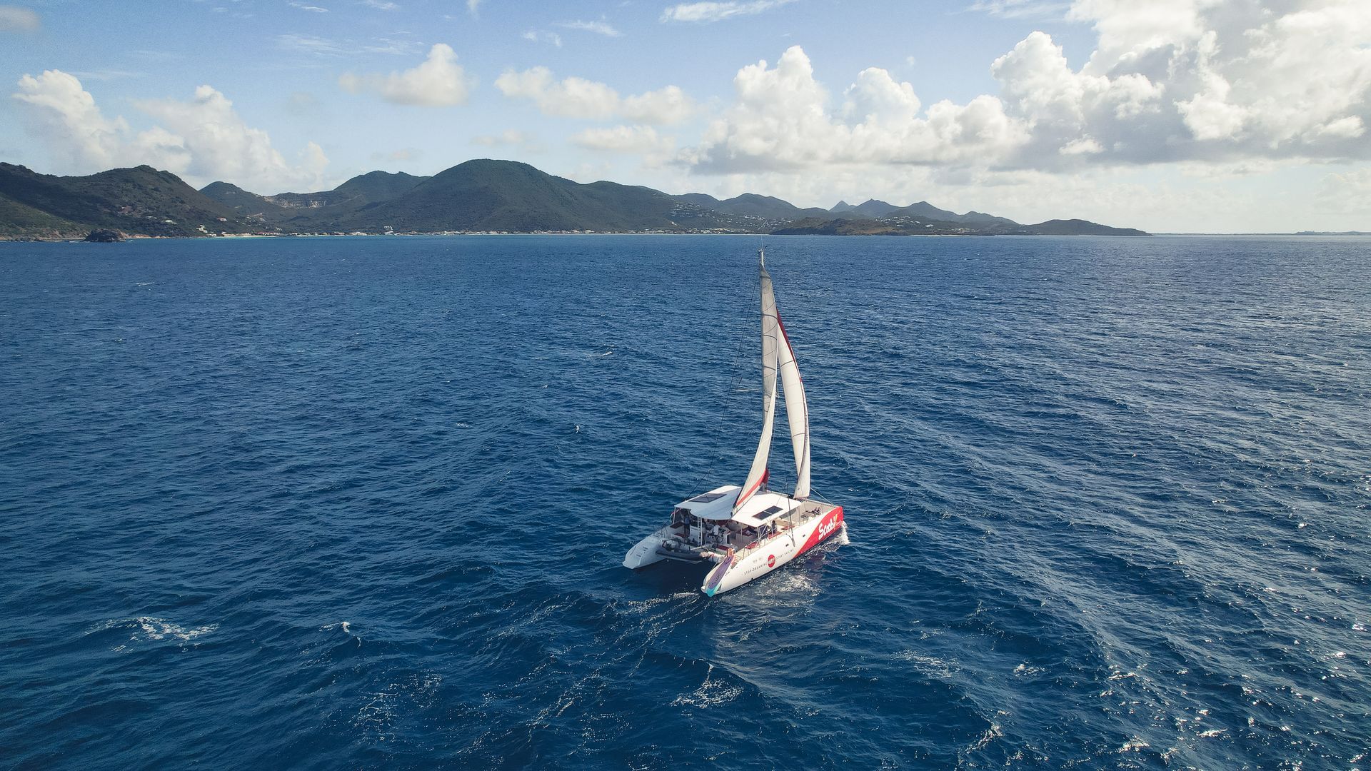 Un catamaran naviguant sur une mer bleue en direction d'une île montagneuse verdoyante sous un ciel nuageux.