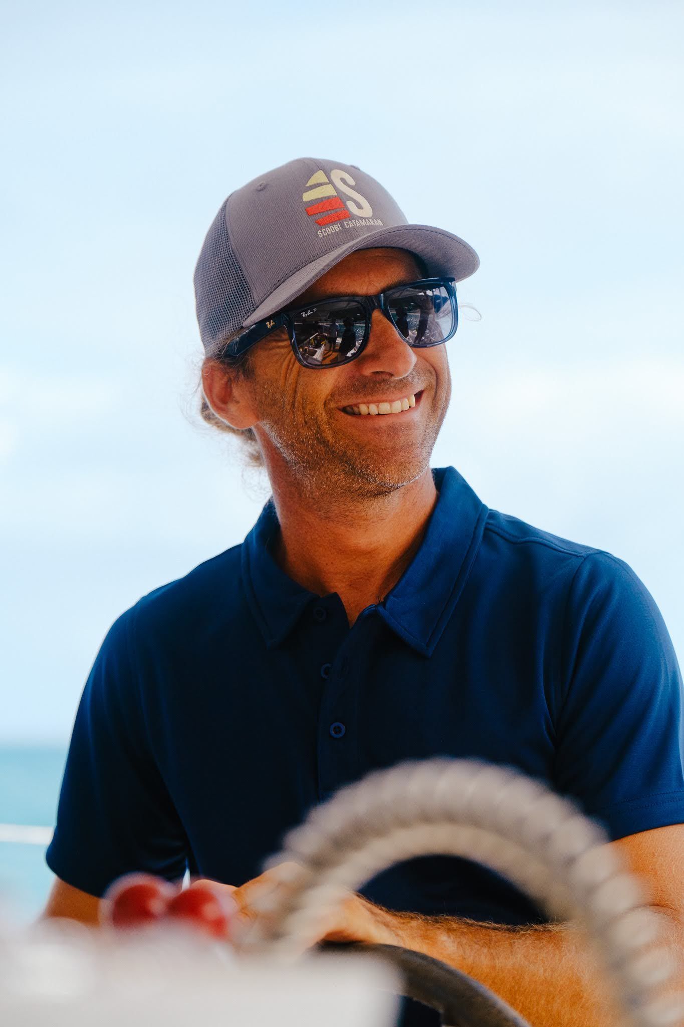 Man steering a sailboat on a sunny day, wearing sunglasses and a hat.
