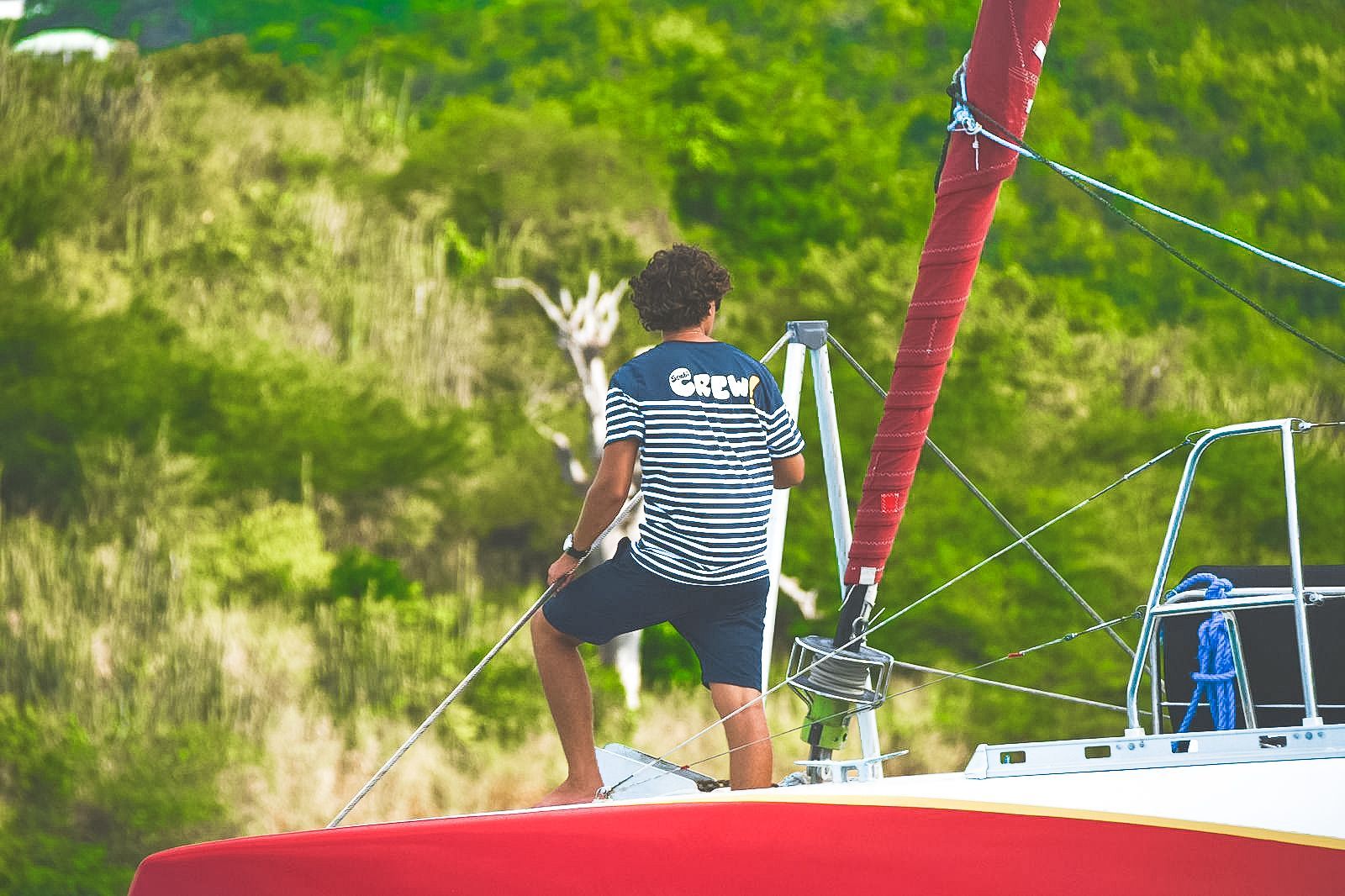 Personne sur un voilier regardant vers une colline verdoyante. Voile rouge, short bleu, chemise rayée.