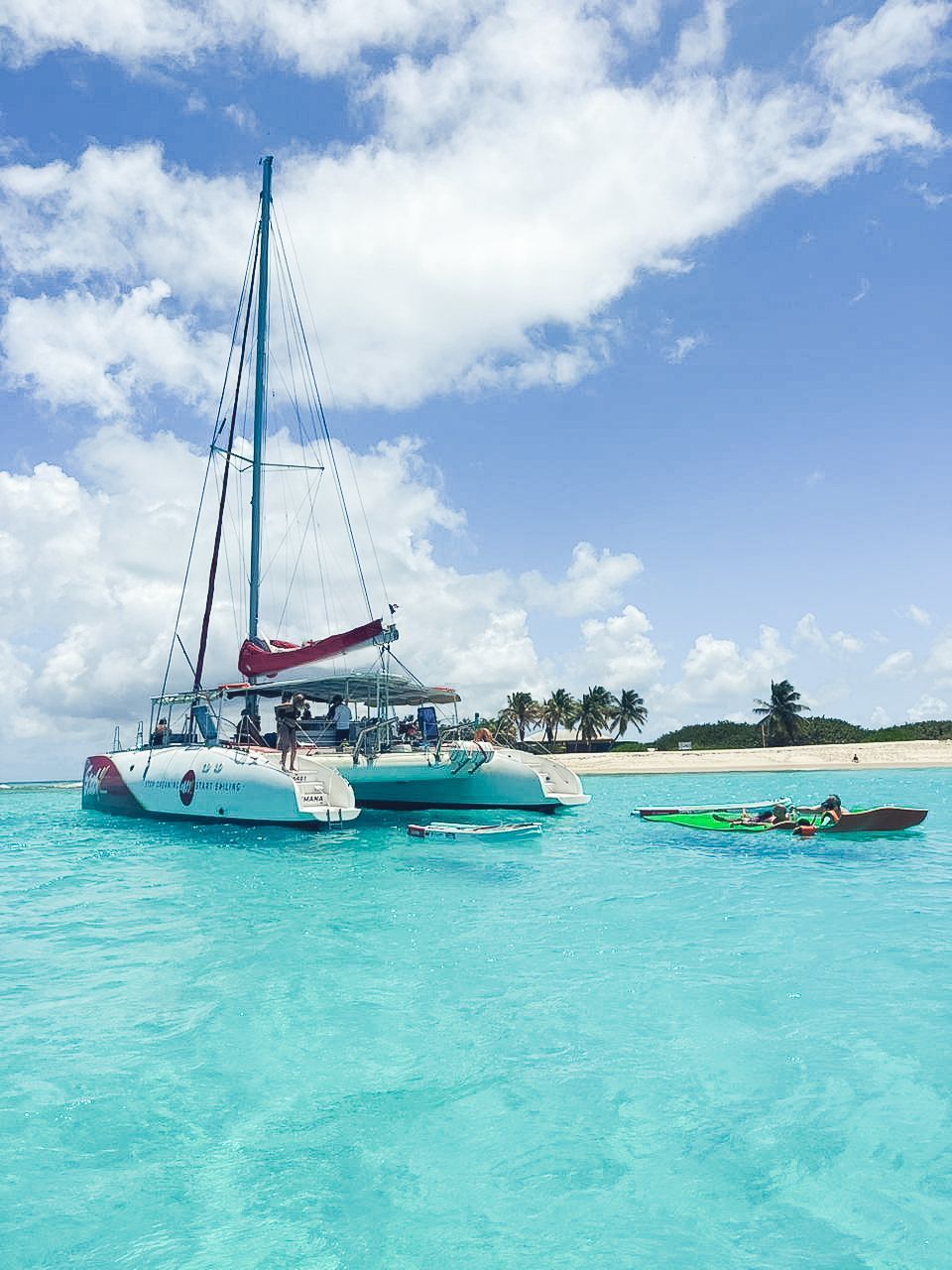 Un catamaran navigue sur des eaux turquoise, avec un petit bateau à proximité. Ciel bleu nuageux et plage en arrière-plan.