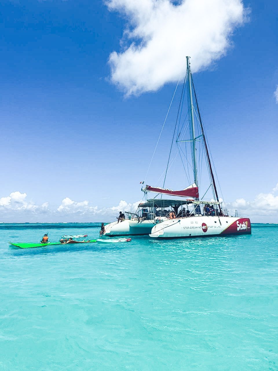 Un catamaran navigue sur une eau turquoise limpide sous un ciel bleu parsemé de nuages ​​; des personnes se baignent à proximité.
