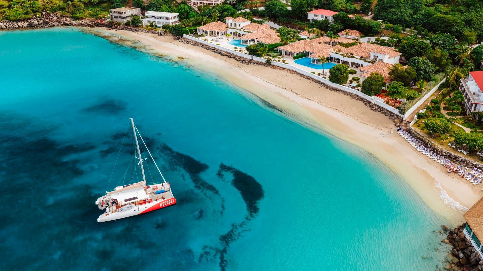 Bright turquoise water, white sand beach, and resort buildings, with a catamaran.