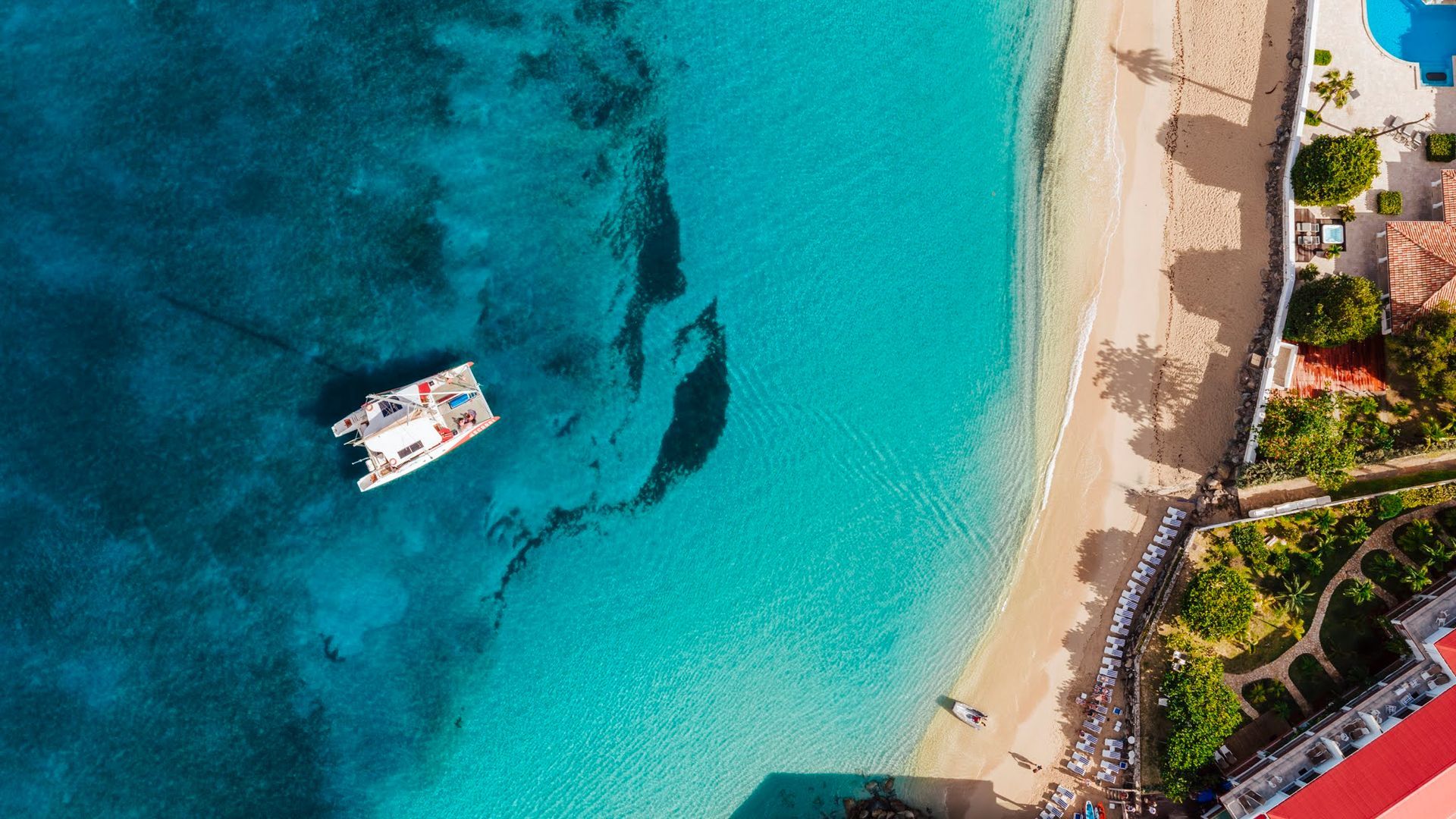 Aerial view of a white boat on turquoise water near a sandy beach with buildings and greenery.