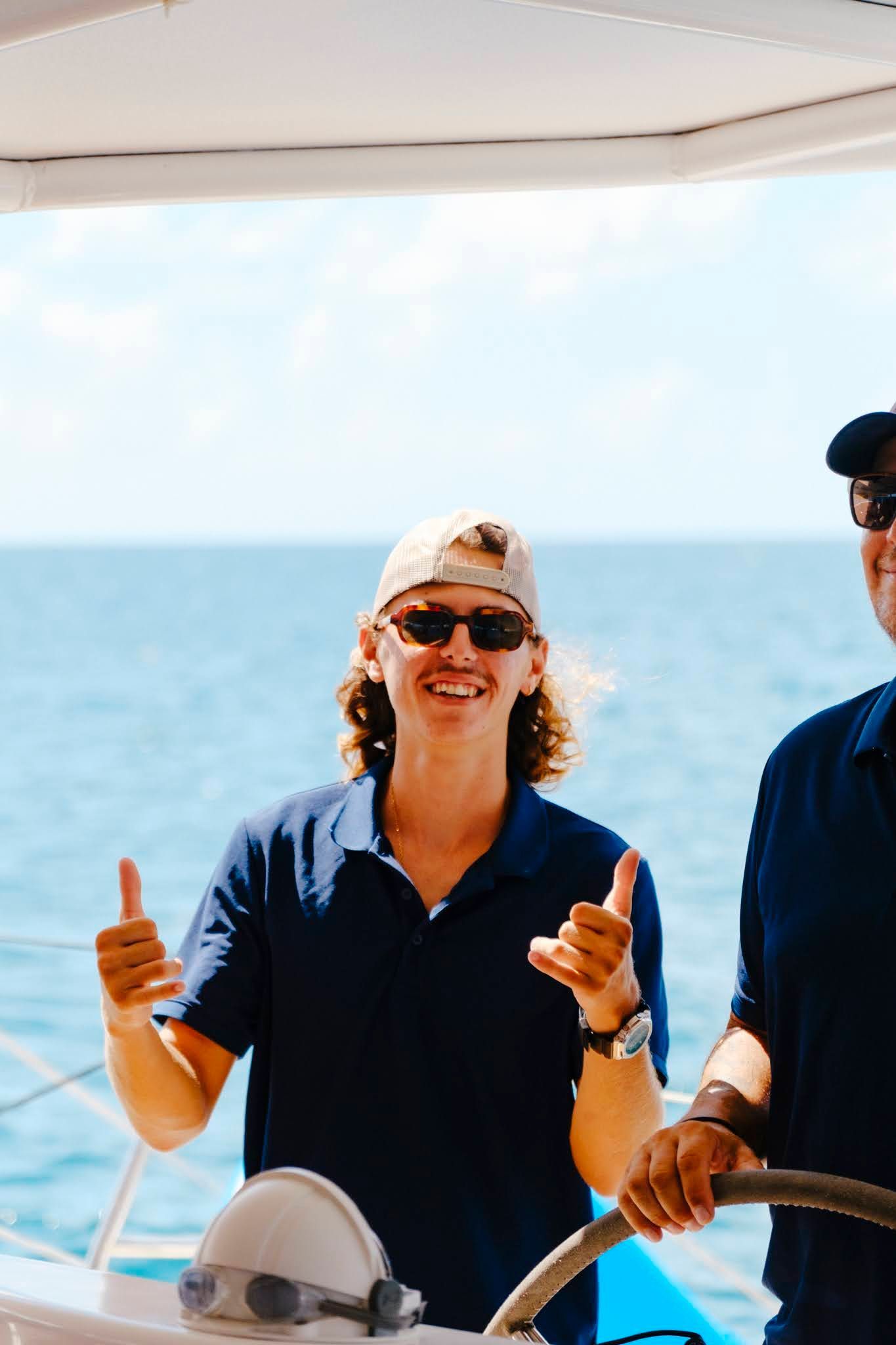 Young man on a boat, giving thumbs up. Smiling, wearing a cap and sunglasses, blue ocean background.