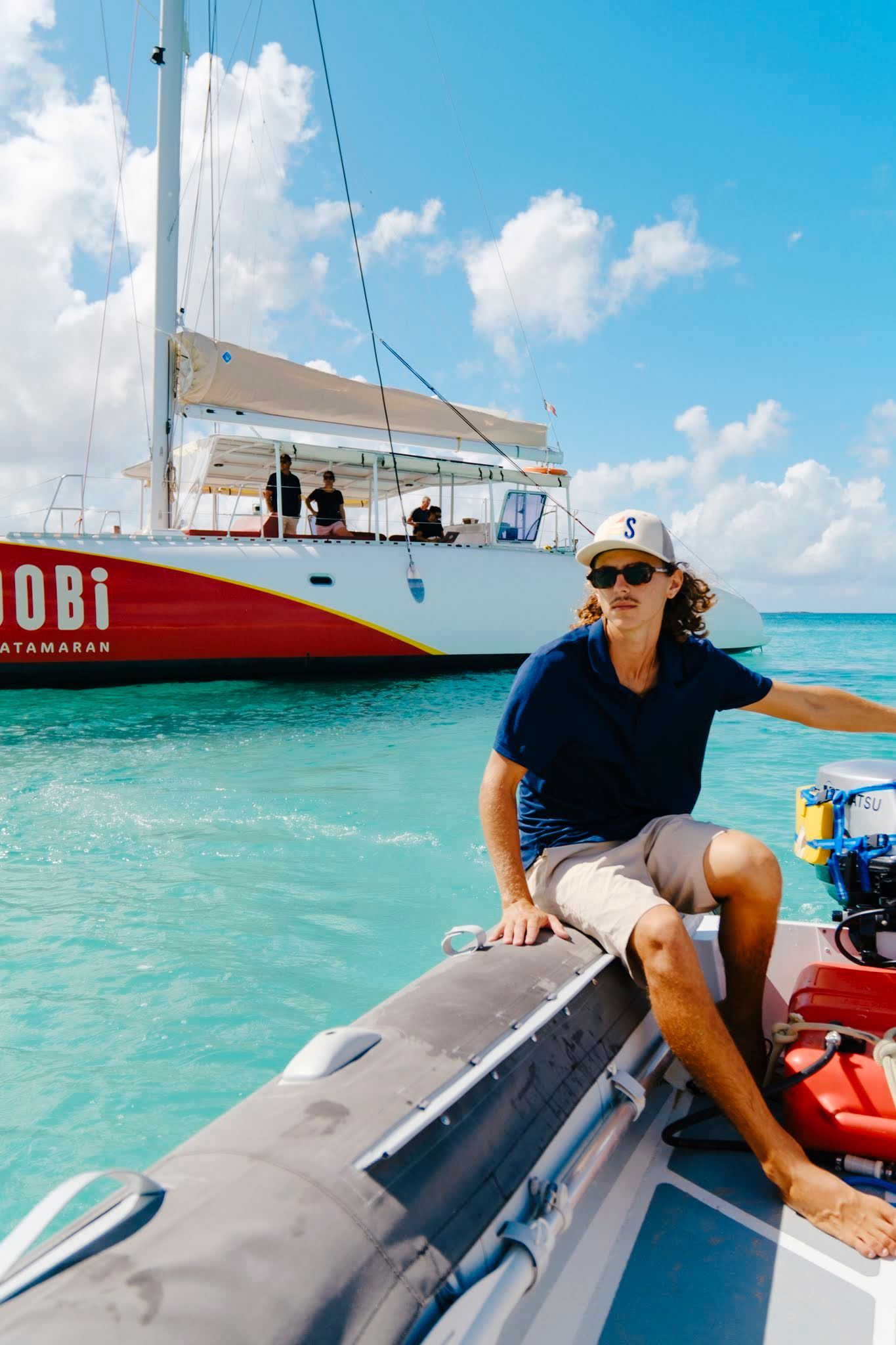 Person on inflatable boat, pointing toward a large sailboat in turquoise water. Bright sunny day.