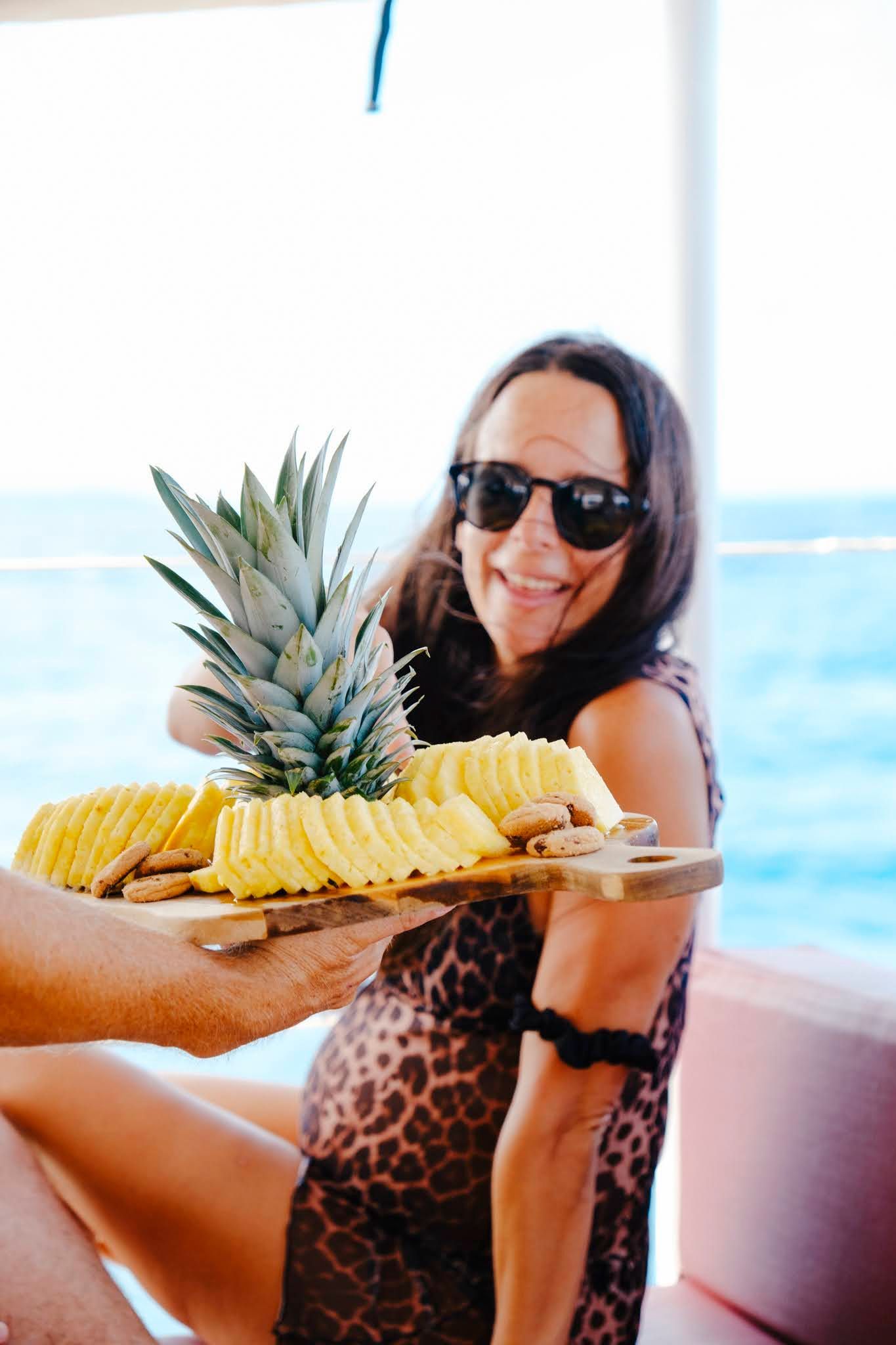 Woman holding a wooden tray of sliced pineapple on a boat, smiling, wearing sunglasses and a leopard-print outfit.