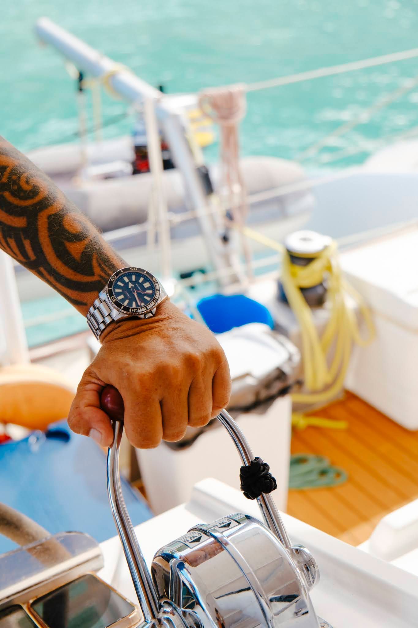 Person's tattooed arm on a boat's throttle. Watch on wrist, ocean background.