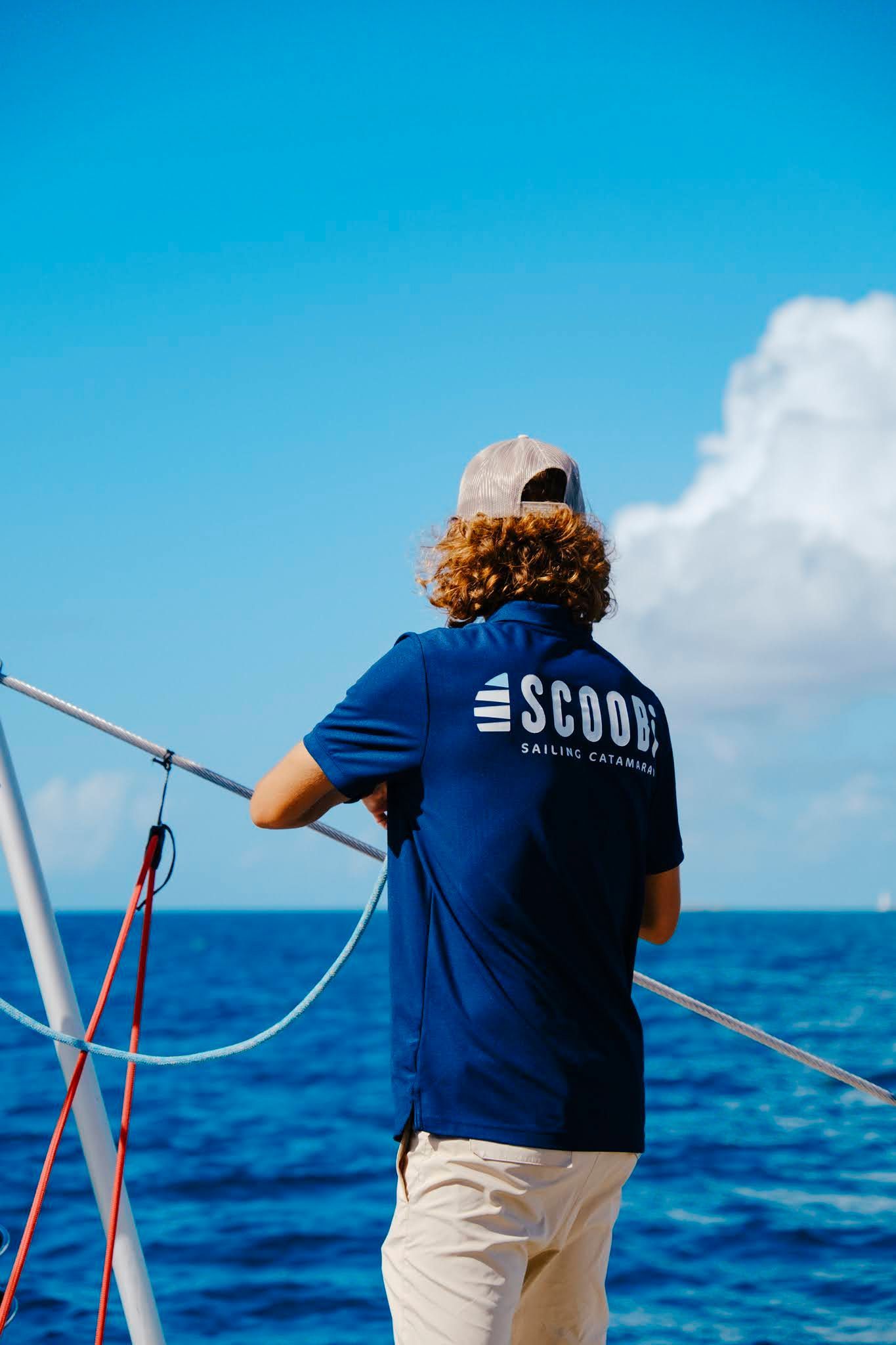 Person on boat, looking at the ocean. Wearing a blue shirt and hat, leaning on a railing. Bright blue sky and water.