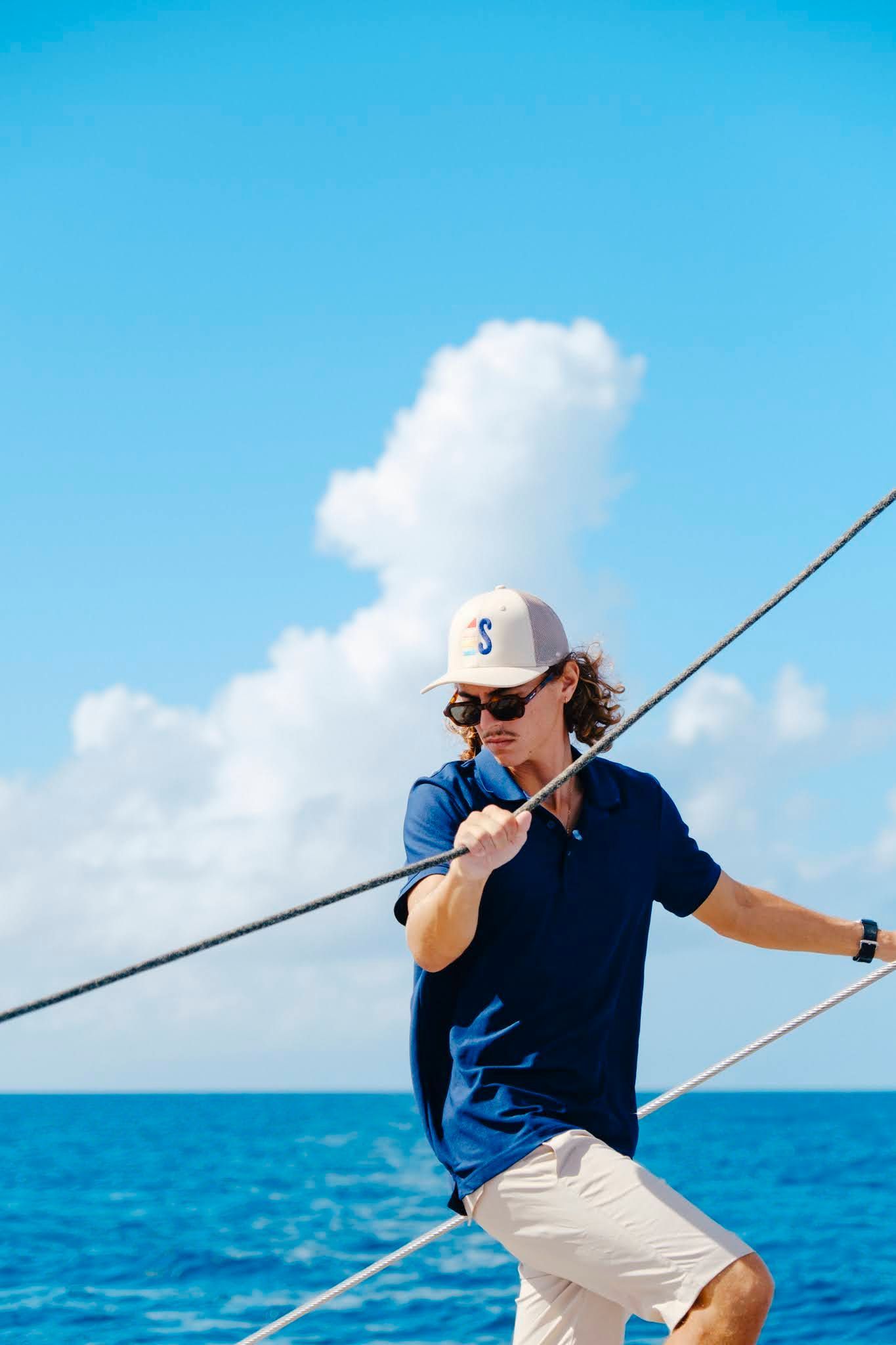 Man on a sailboat, holding a rope, blue shirt, tan hat, sunny day on the ocean.