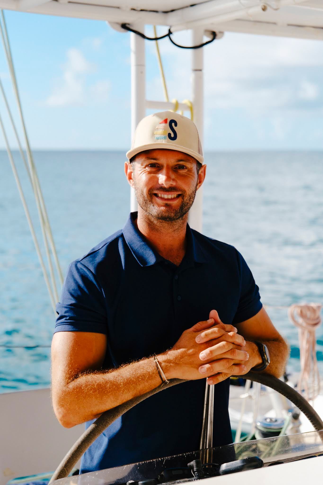 Man at the helm of a sailboat, smiling. He wears a blue shirt and cap; ocean visible.