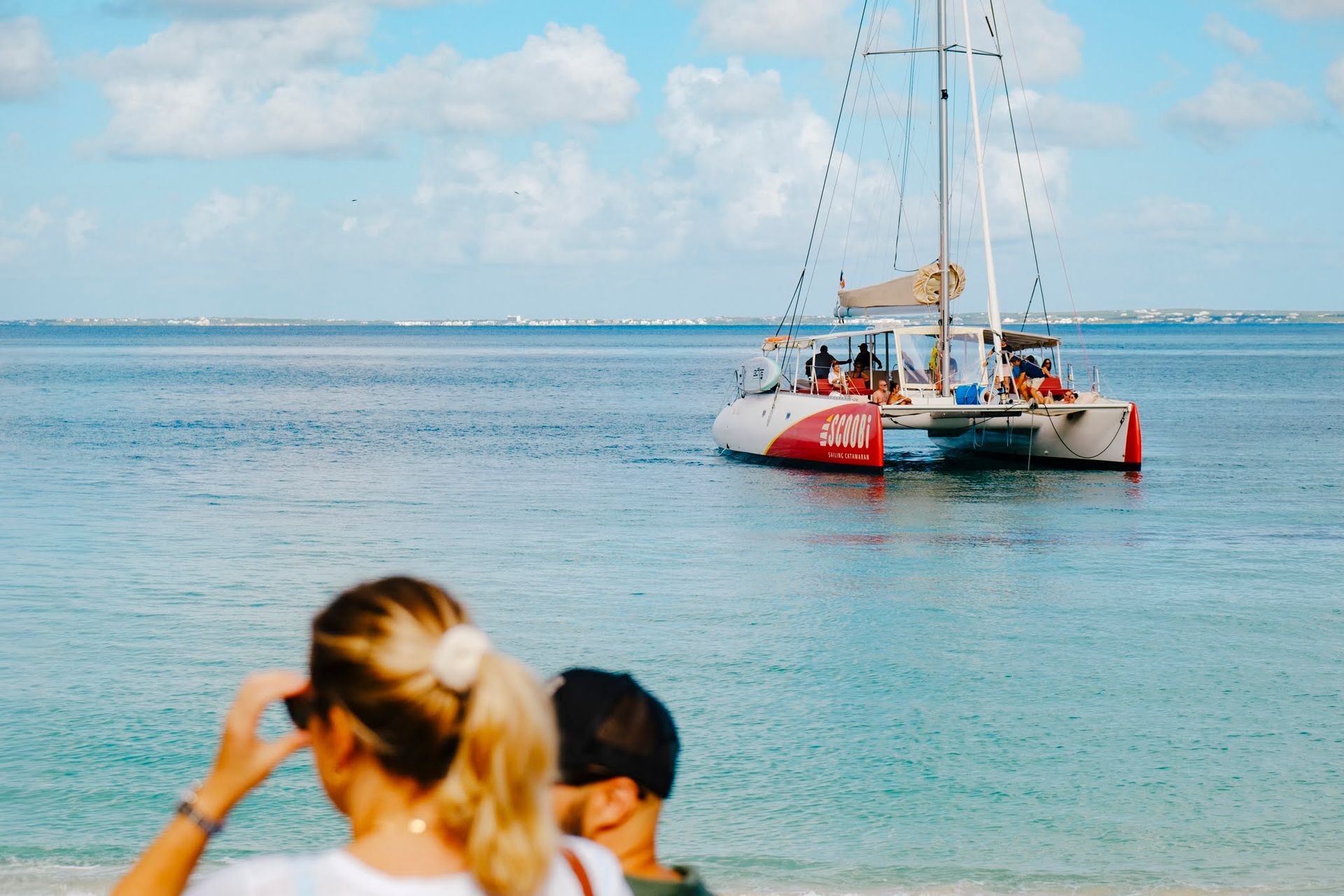 People watch a catamaran on the ocean. The boat is red and white with a beige sail. Blue sky and water.