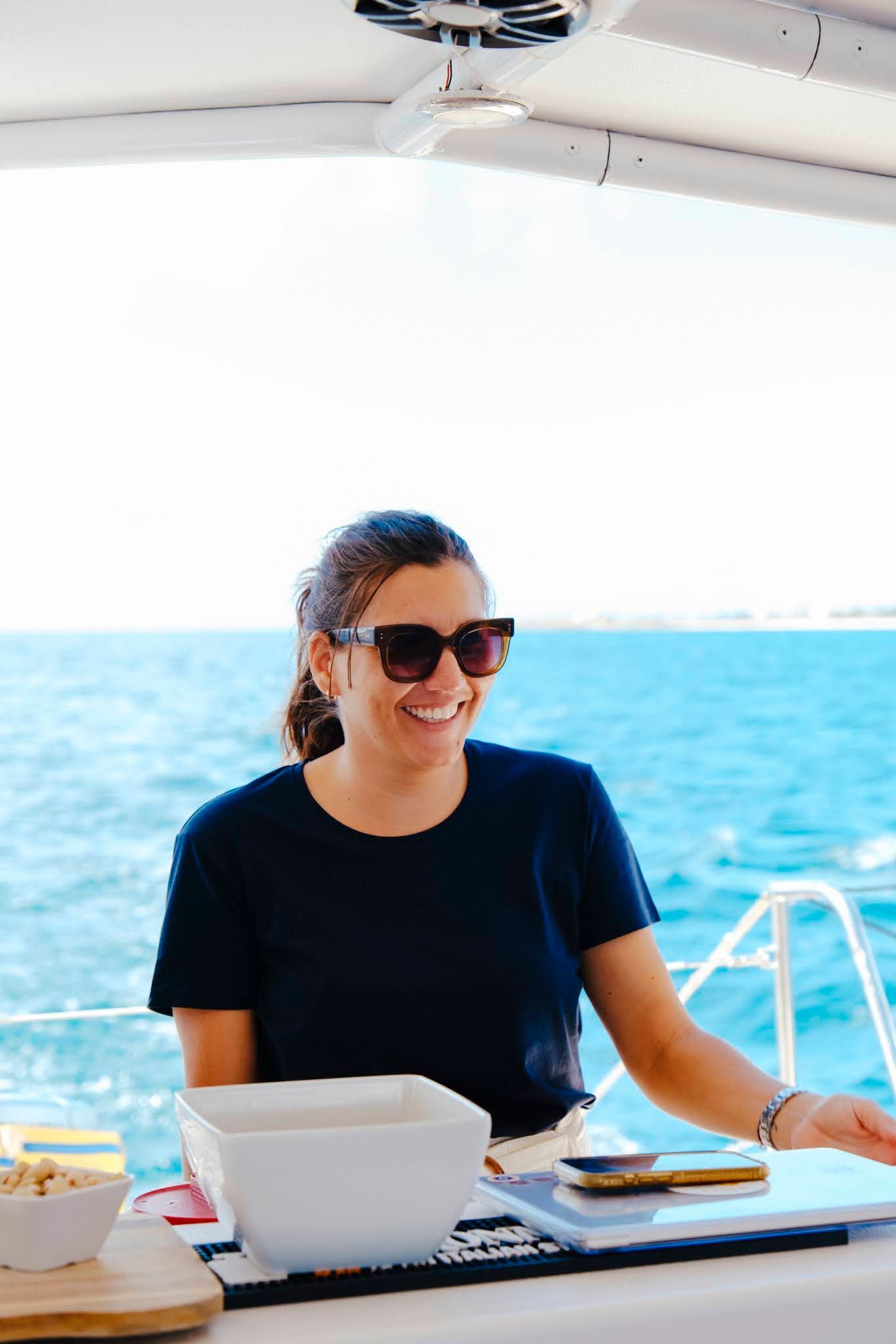 Femme souriante sur un bateau, portant des lunettes de soleil, mer bleue en arrière-plan.