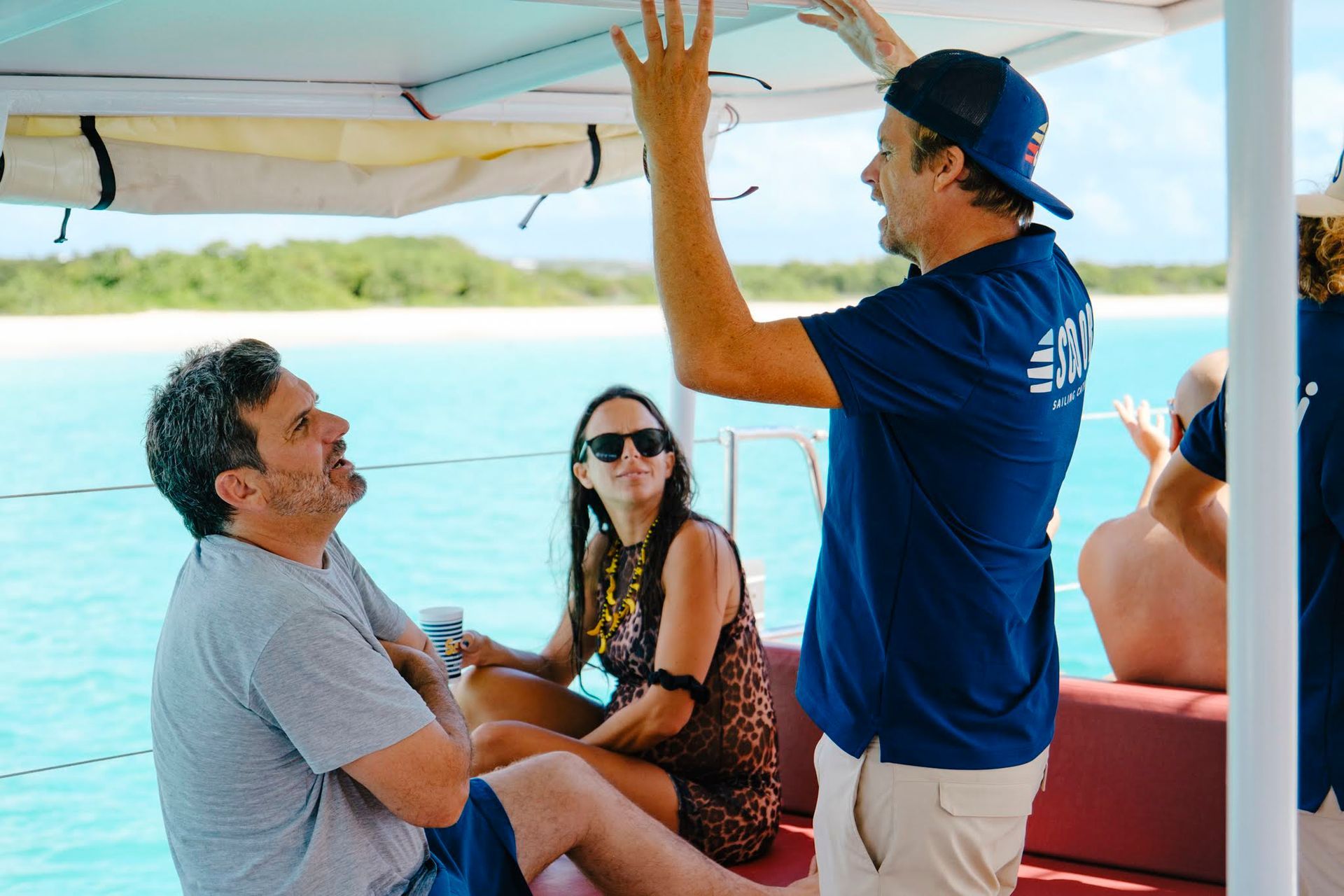 People on a boat with turquoise water; one man gestures, two others seated smiling, sunny day.