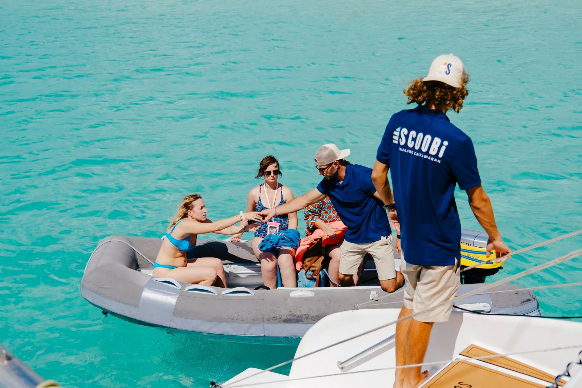 Des personnes se tapent dans la main à bord d'une petite embarcation tirée par un plus grand bateau, en pleine mer. L'homme en chemise bleue tire sur la corde.