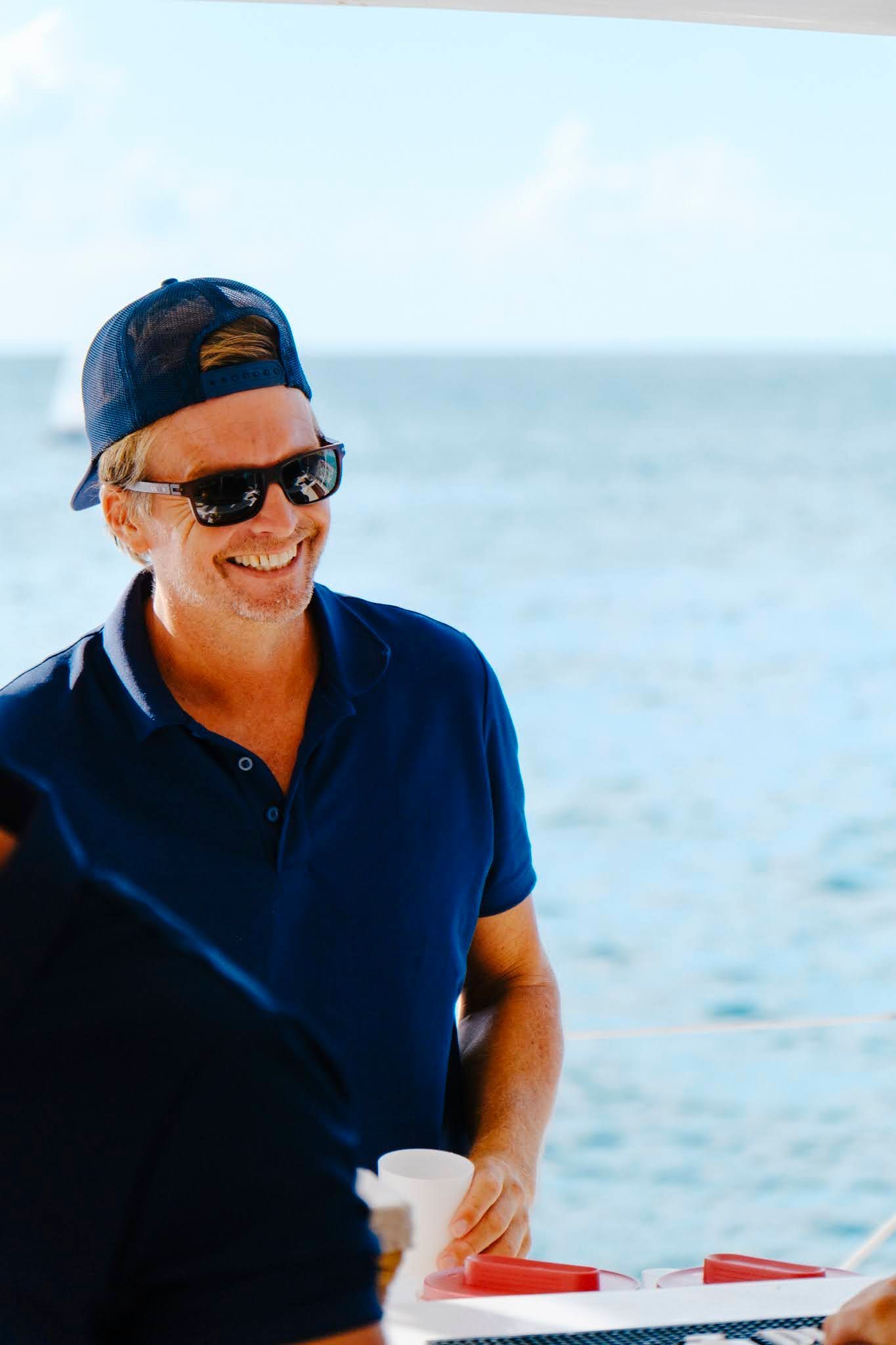 Man wearing a blue cap and sunglasses, smiling on a boat with the ocean in the background.