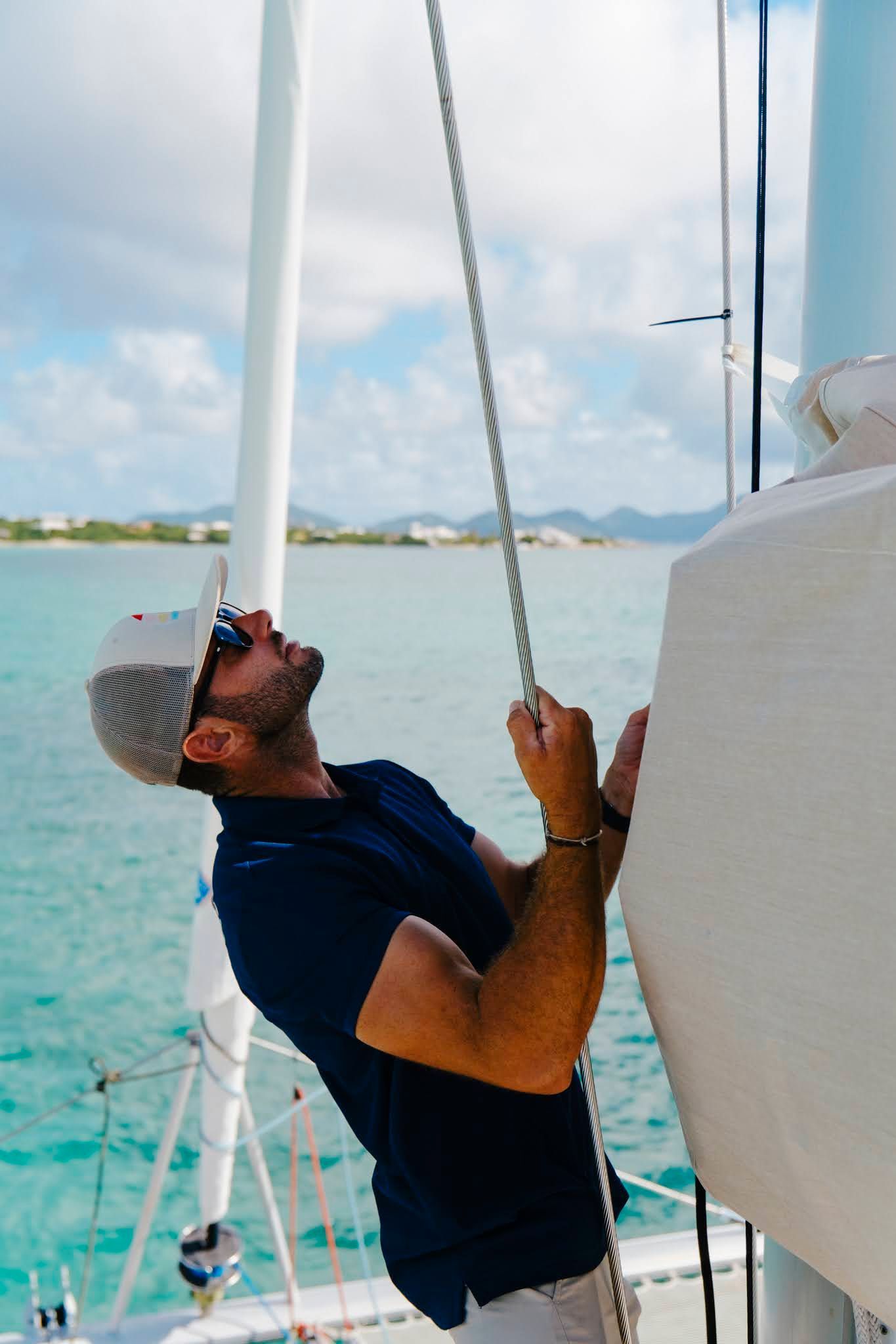 Man on sailboat, pulling rope up mast, looking up at sail, wearing hat and sunglasses.