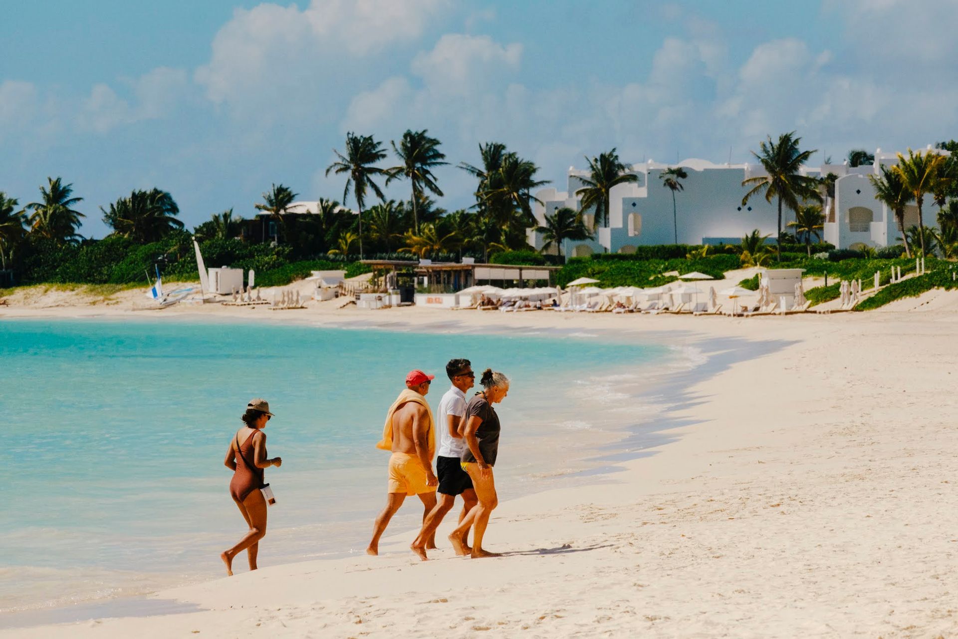 People walking on a white sandy beach with turquoise water, palm trees, and white buildings in the background.