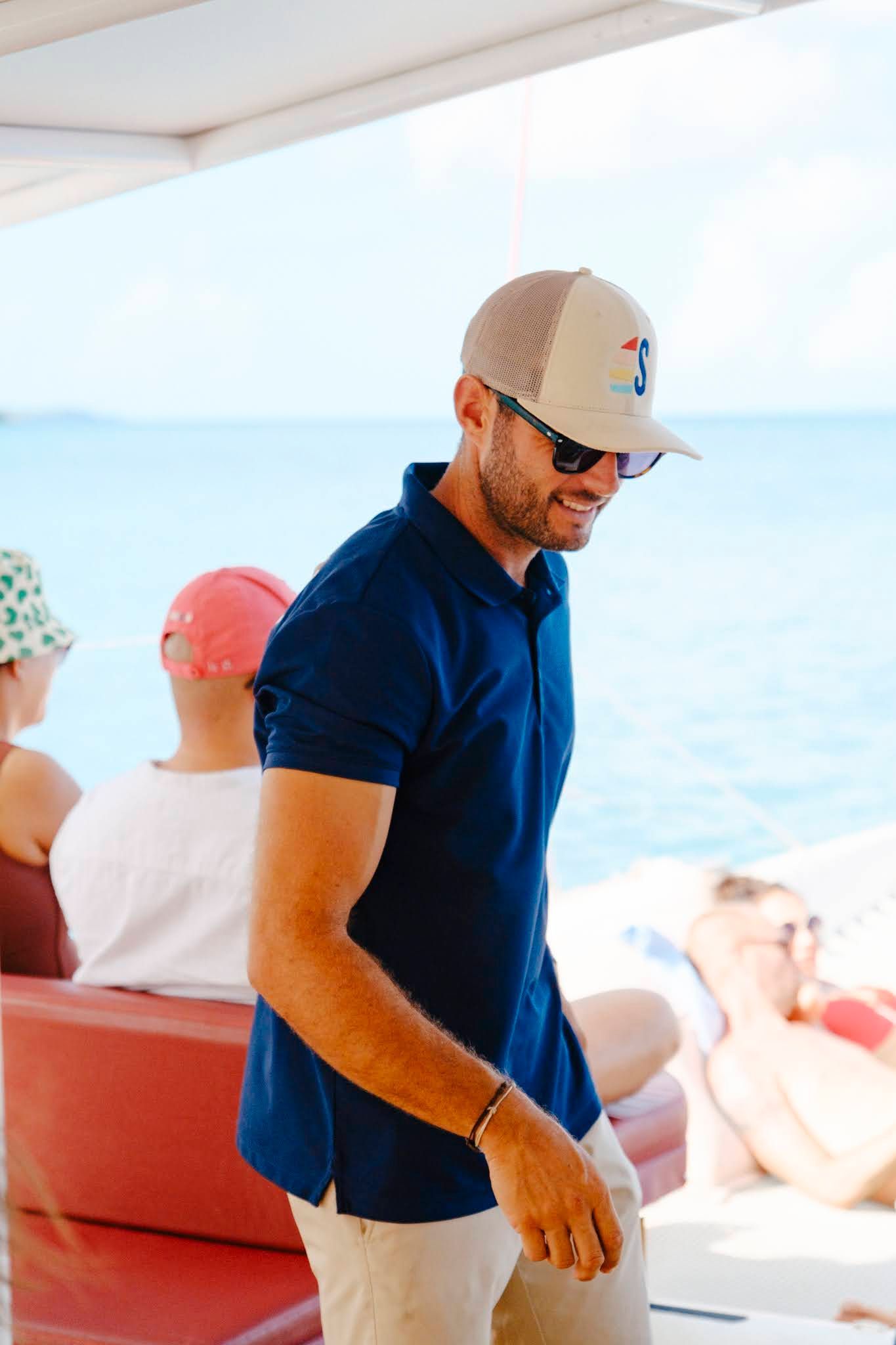 Man smiling, wearing a blue polo shirt, tan hat, and sunglasses on a boat. Ocean background.