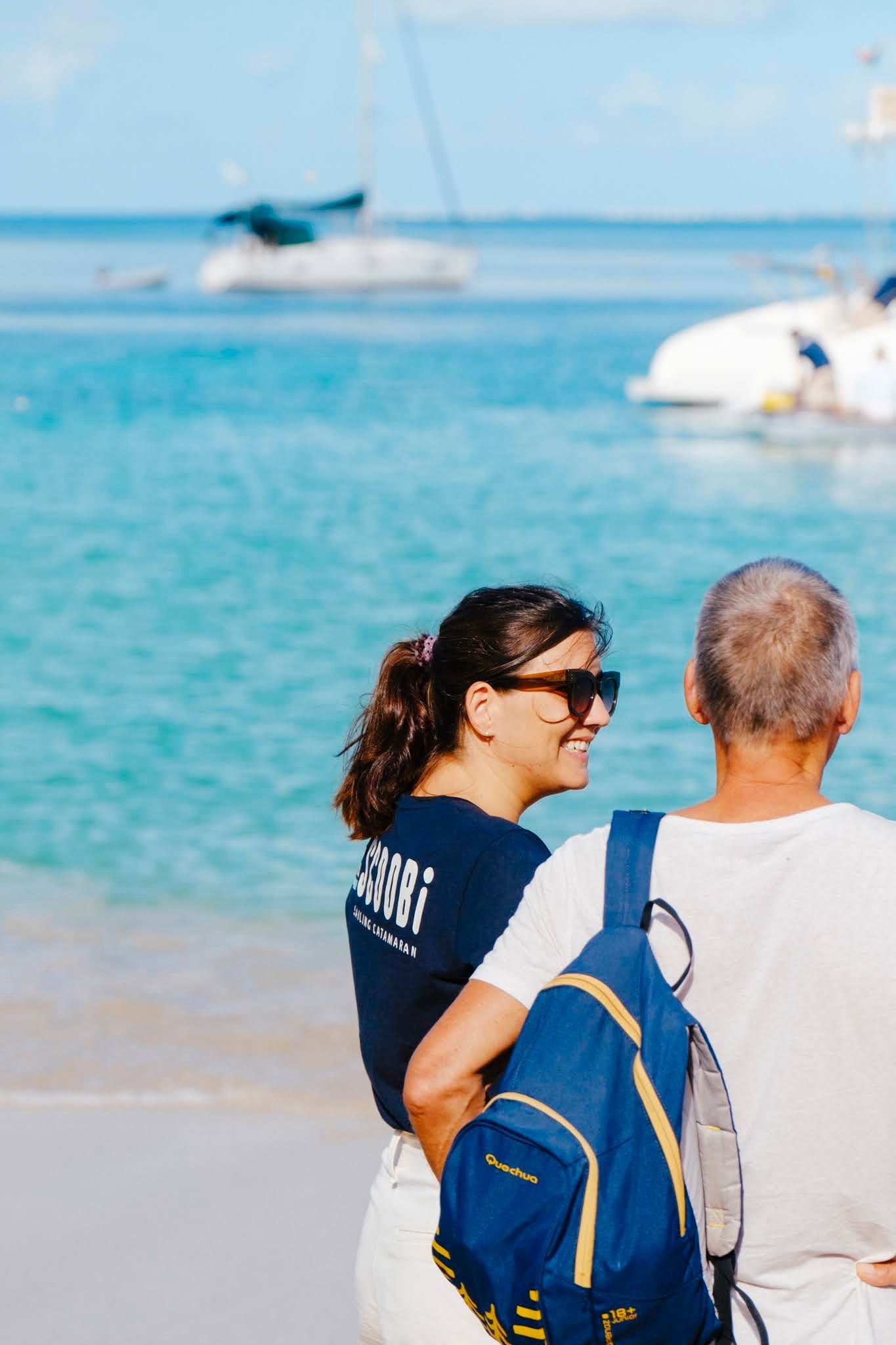 Woman in sunglasses smiles at a man on a beach; boats in the background, blue water.