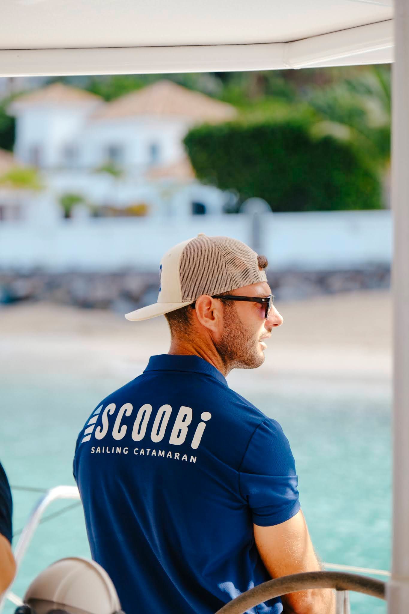 Man in blue shirt and cap on a boat, looking toward a waterfront building.