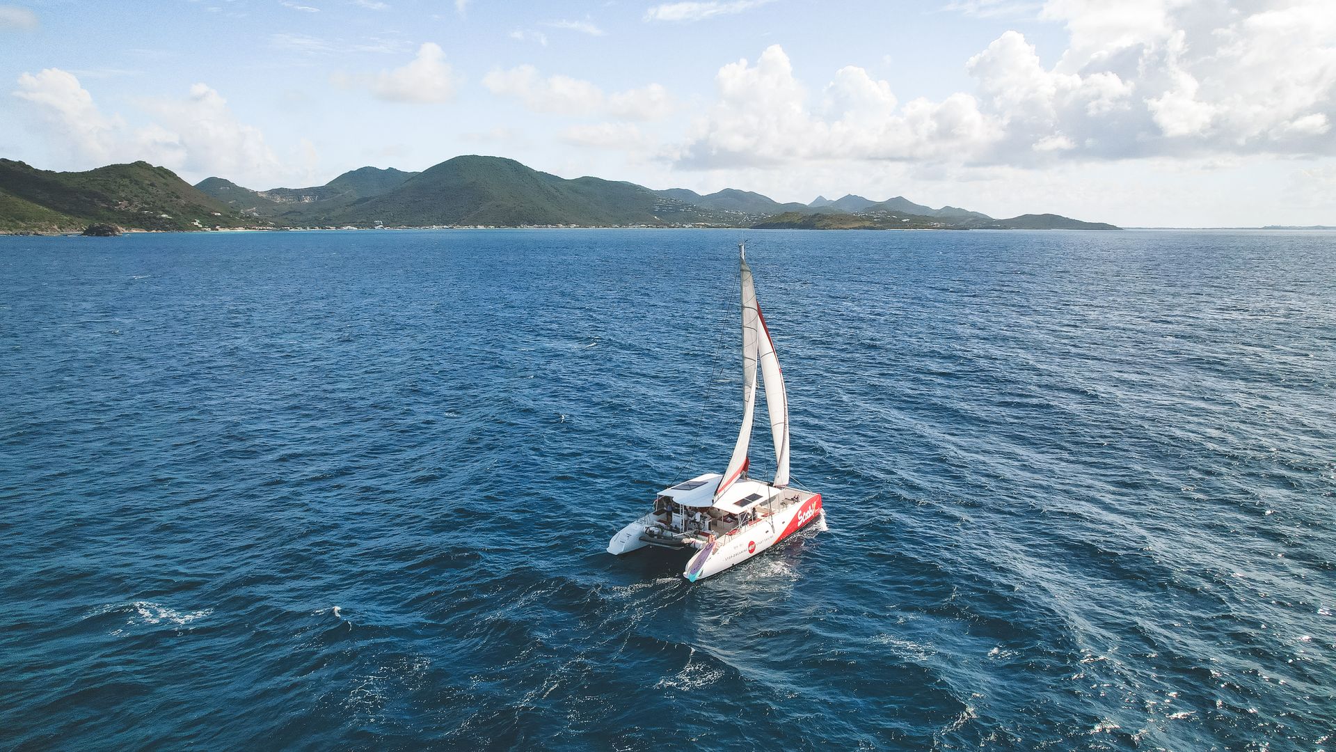 Catamaran sails on blue ocean, hills in background.