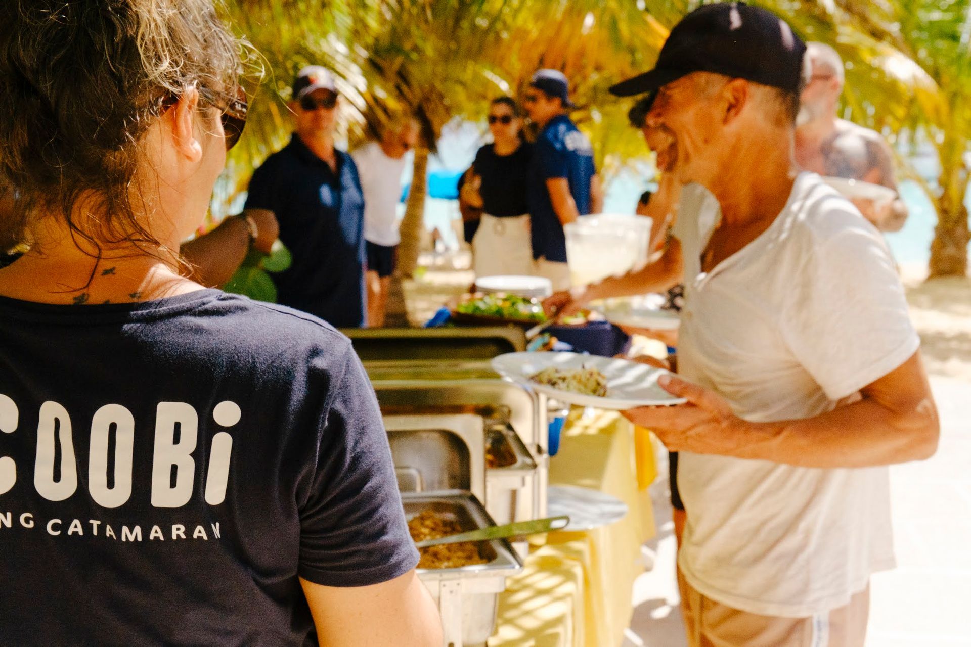 Des personnes attablées à un buffet de plage, servies par un serveur portant un t-shirt « Coobi ». D'autres personnes sont visibles en arrière-plan.