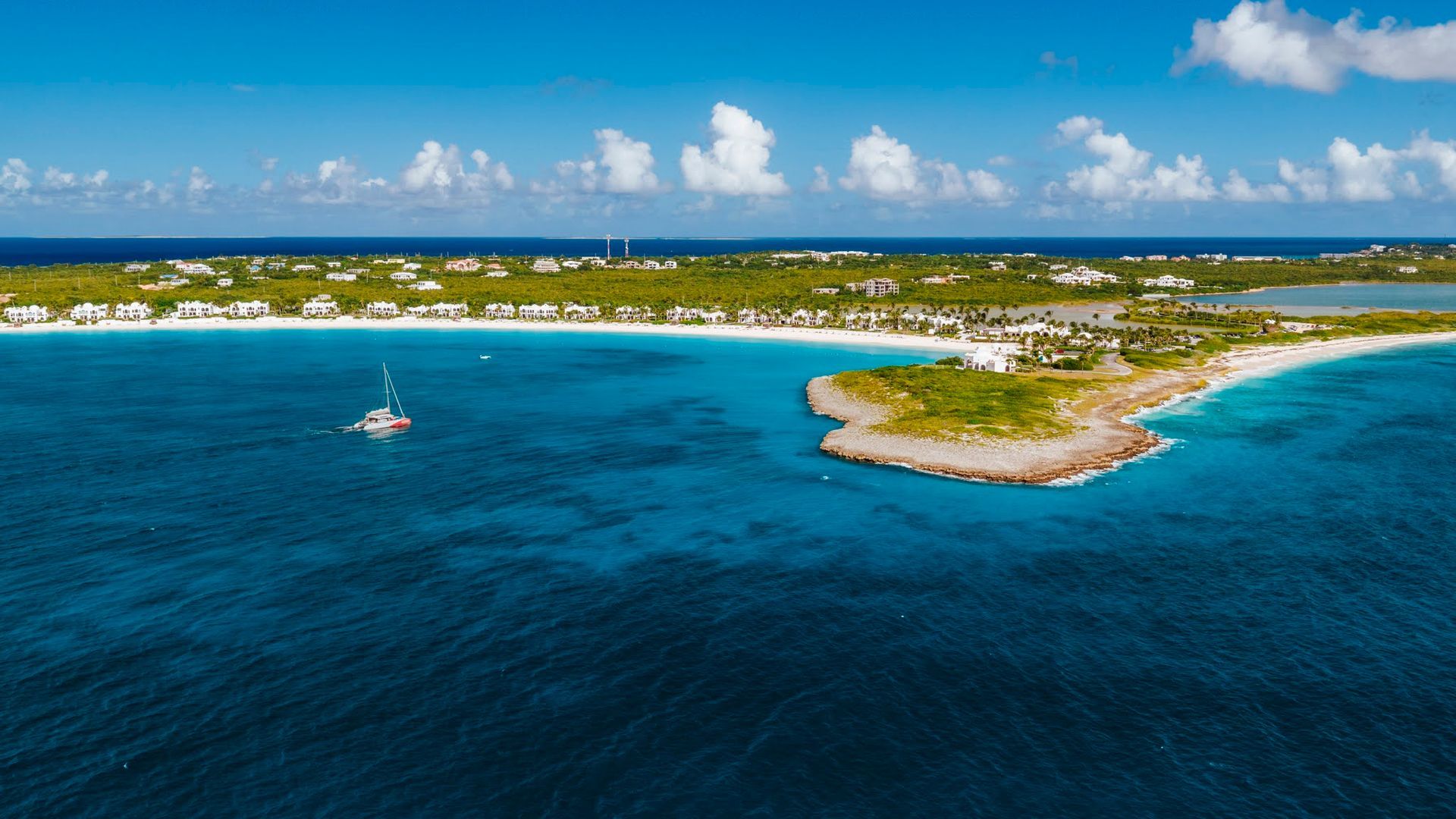 Vue aérienne d'une plage de sable blanc, d'une eau turquoise et d'une petite île sous un ciel bleu.